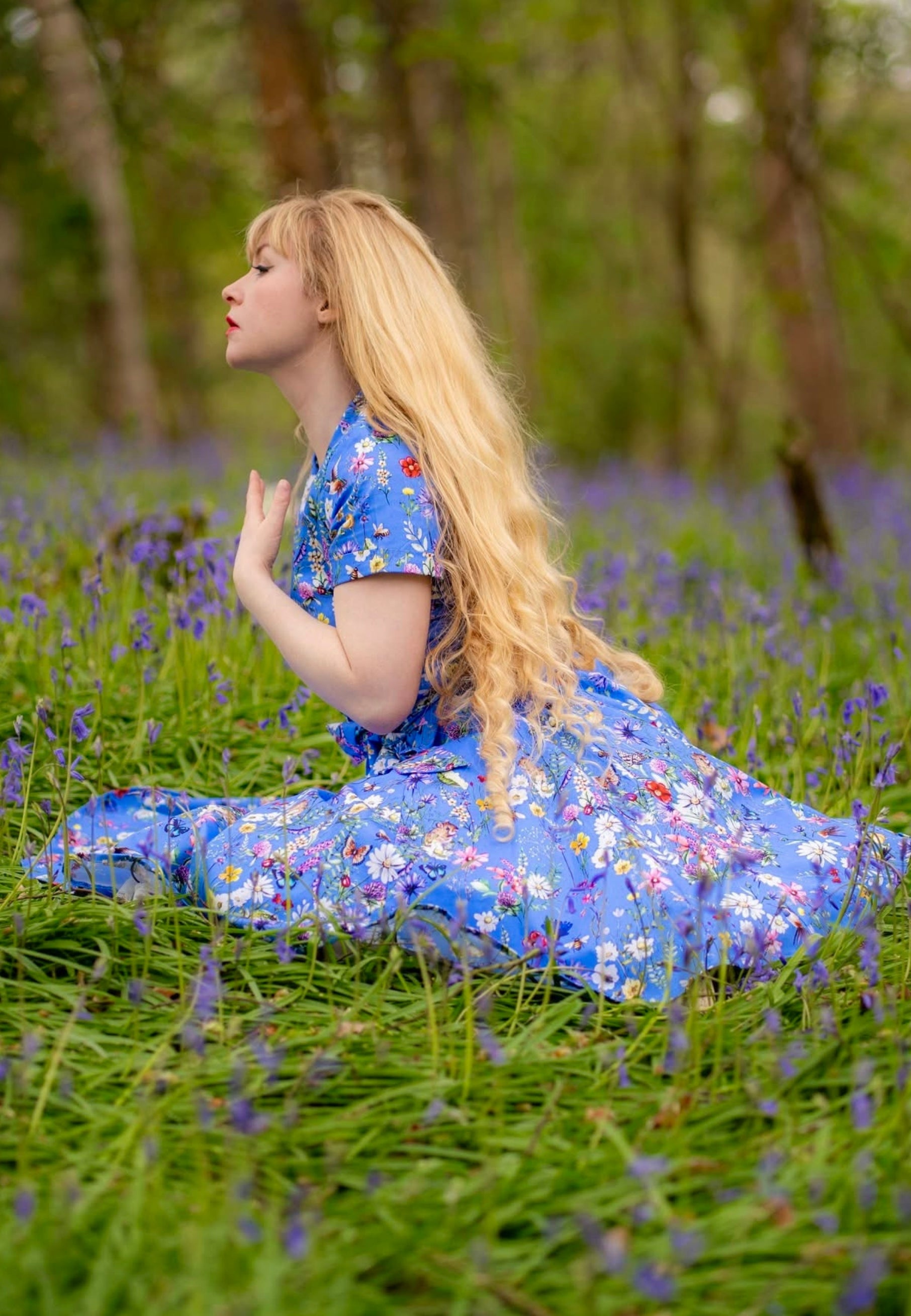 Woman with long blonde hair sat in a wild flower area of a wood wearing a beautiful cornflower blue dress with a wild flower print. 