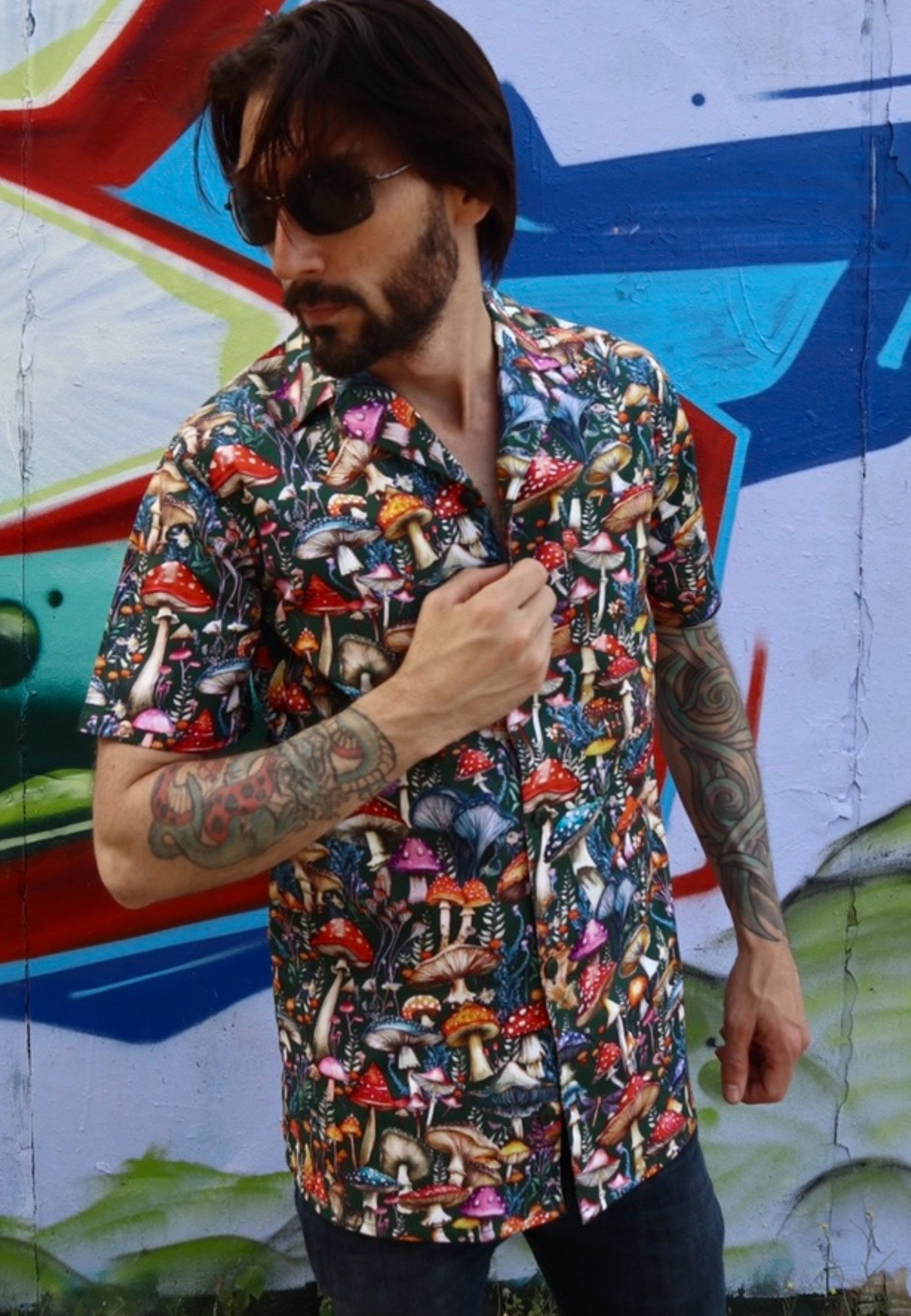 Man wearing a colorful mushroom-patterned shirt in front of a graffiti wall.