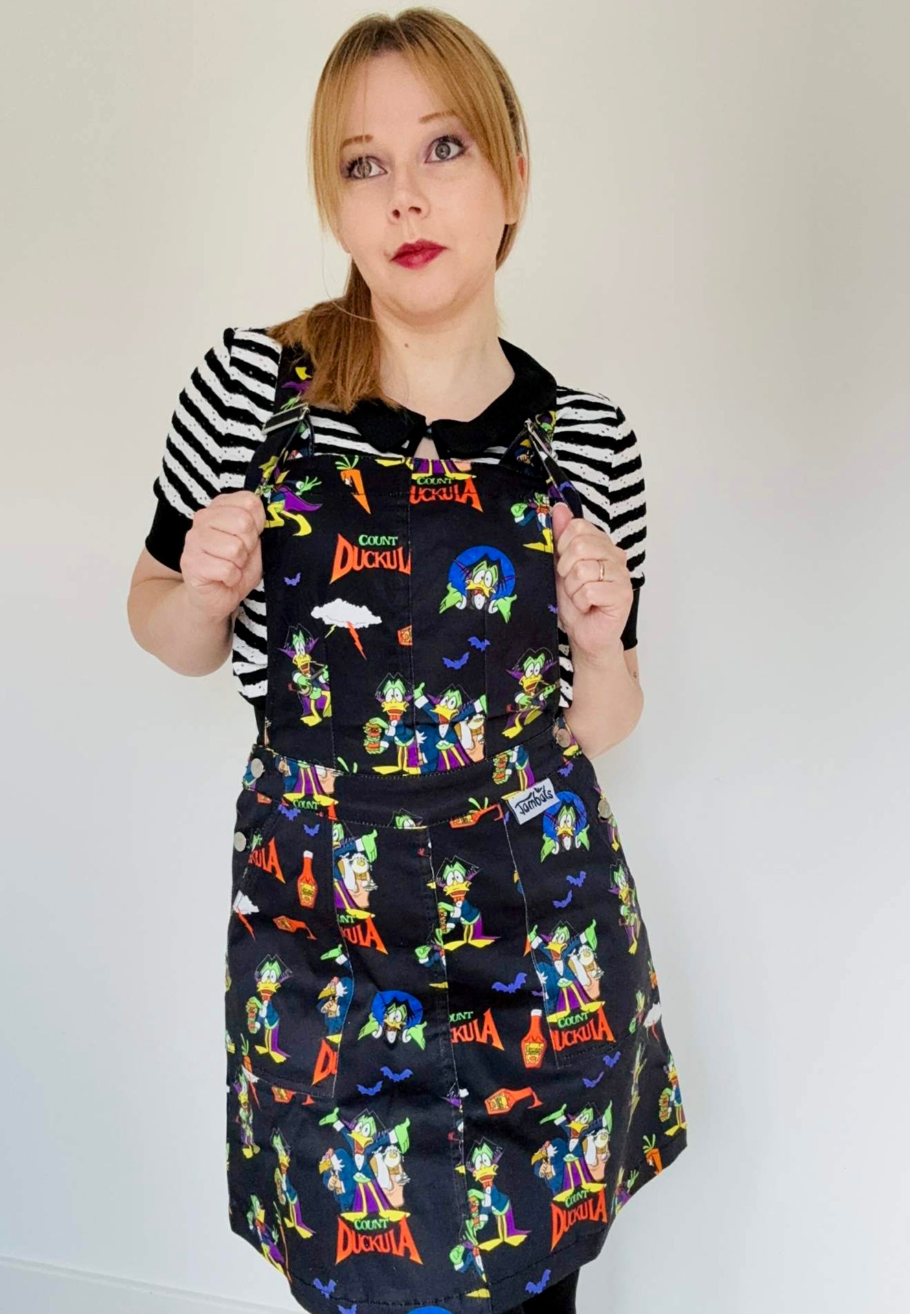 Woman wearing a black pinafore dress with vibrant Count Duckula print. Studio photo. 
