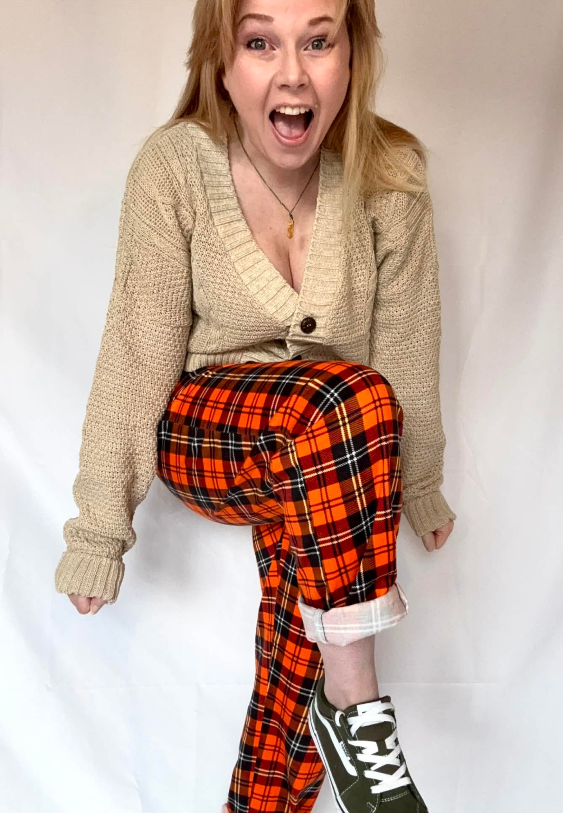 Woman wearing a beige cardigan and orange tartan trousers and black Vans pumps on a white background