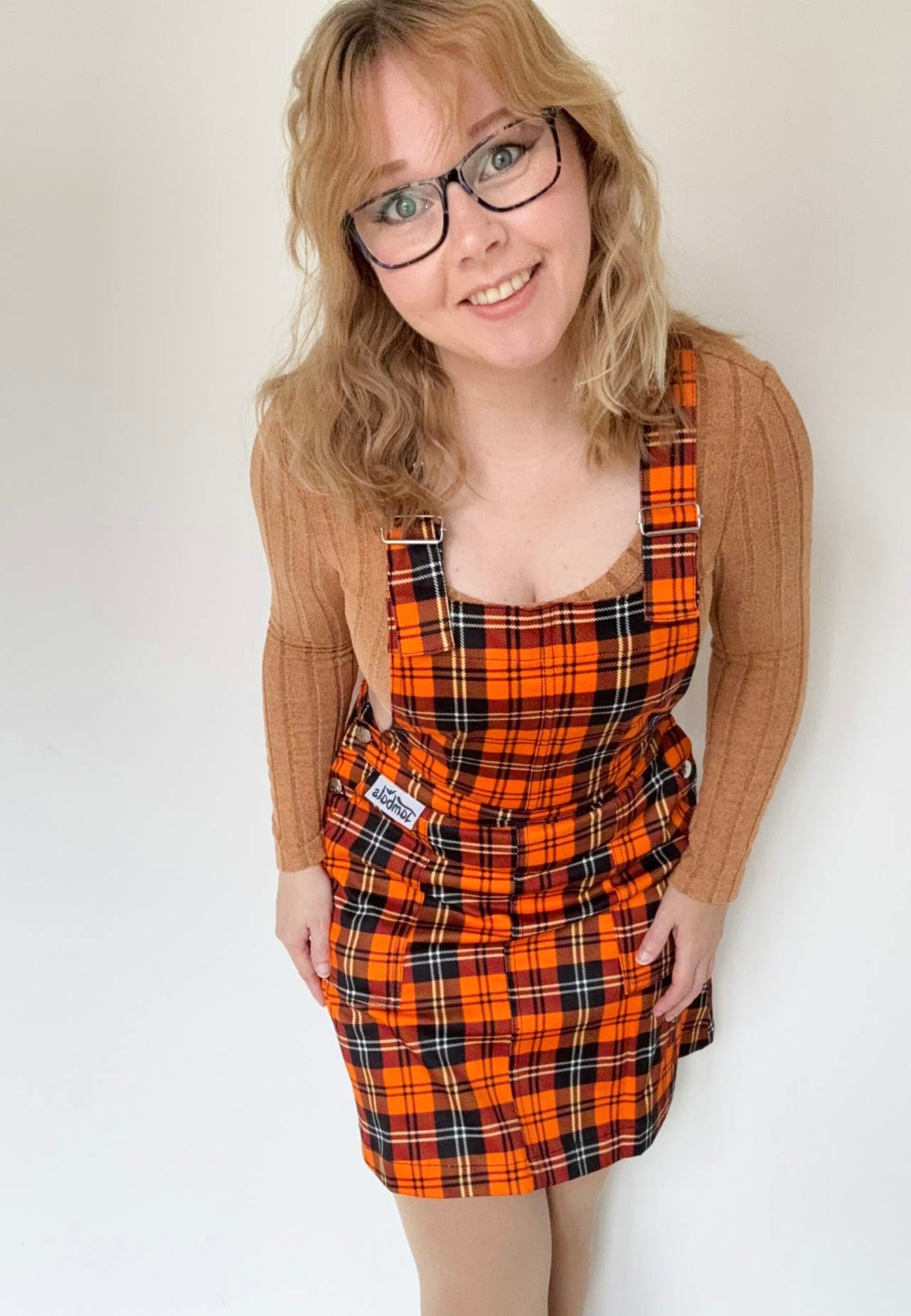 Woman wearing an orange tartan pinafore dress over a sweater against a white background.