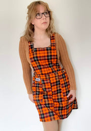Woman wearing an orange tartan pinafore dress over a sweater against a white background. Woman is wearing bookish glasses.