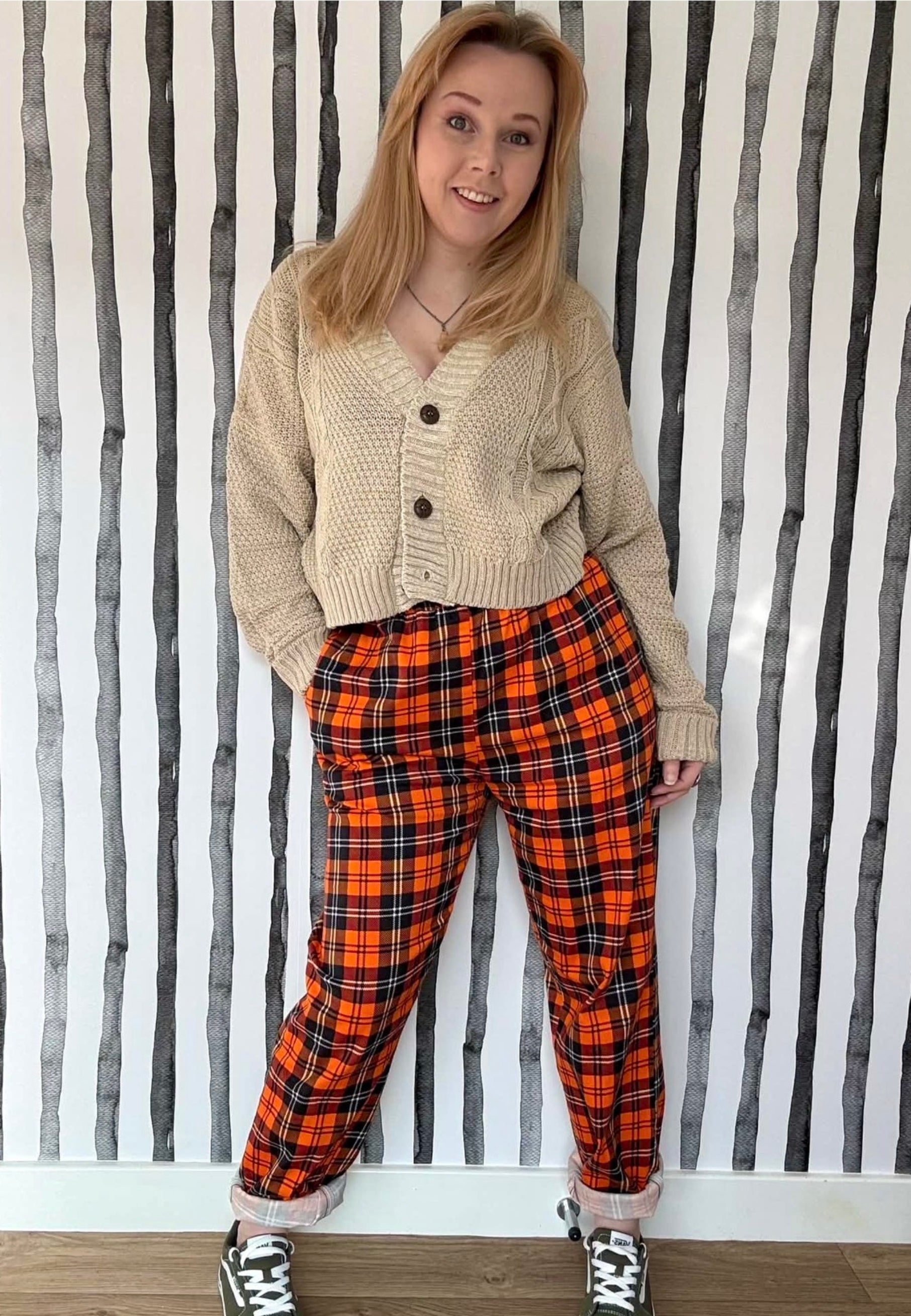 Person wearing a beige cardigan and orange tartan trousers against a striped wall.
