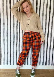 A woman wearing a beige cardigan and orange tartan trousers against a striped wall.