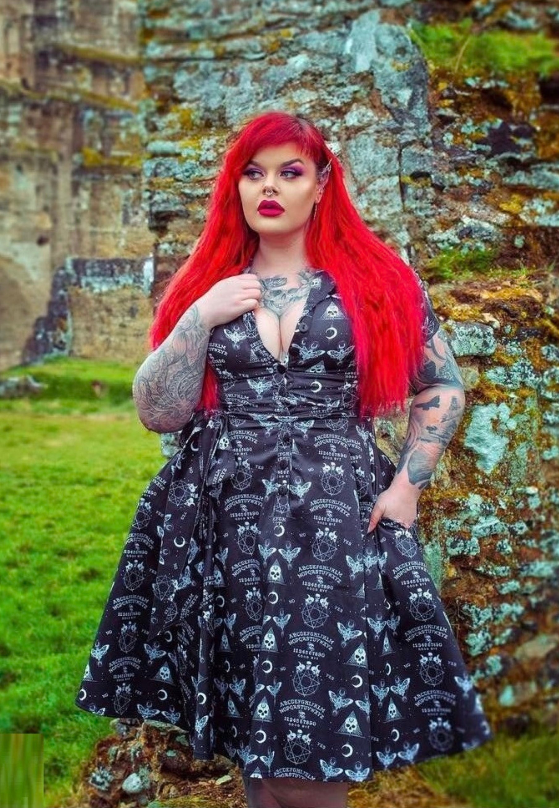 A woman with red hair wearing a black gothic Ouija print dress with a full skirt and button shirt bodice, standing in front of a rock wall.
