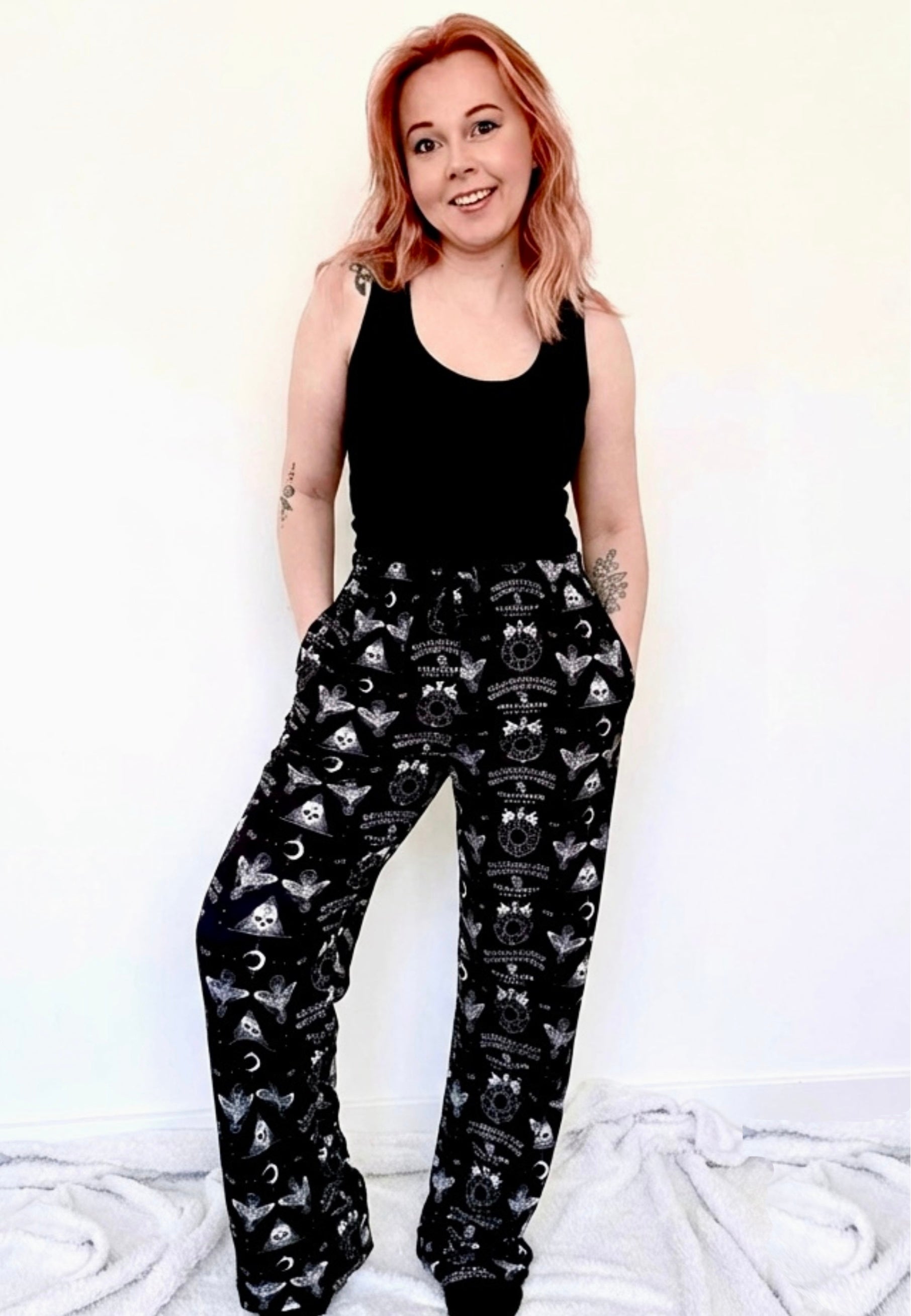 Woman wearing a black sleeveless top and black gothic patterned Ouija print lounge pants on a white background