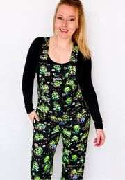 Woman wearing a black long-sleeve shirt and black dungarees overalls with a pattern of cute hippy peas at a music festival of pease and love. Woman is standing against a white background. 