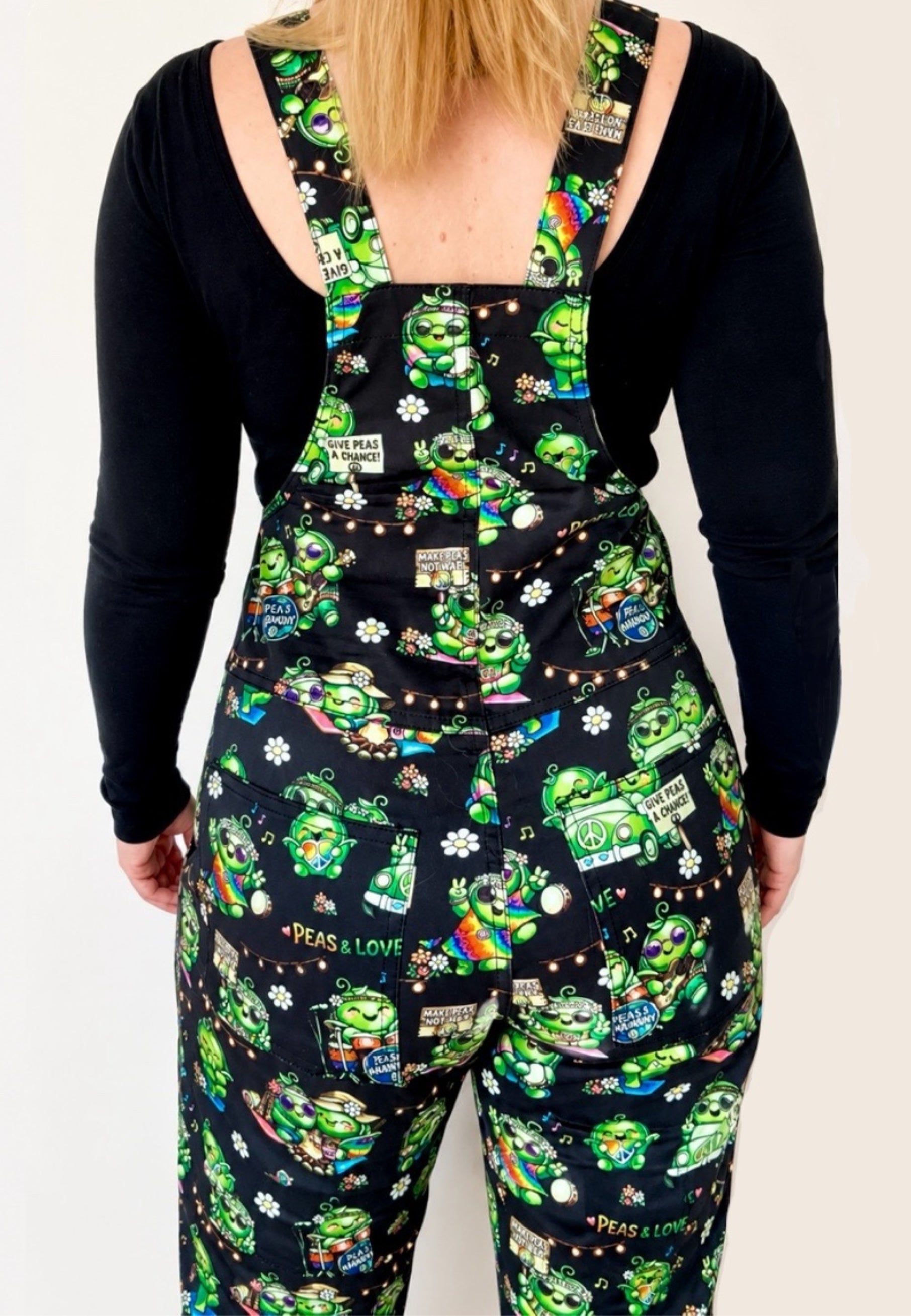 Woman wearing a black long-sleeve shirt and black dungarees overalls with a pattern of cute hippy peas at a music festival of pease and love. Woman is standing against a white background. Back view of dungarees. 