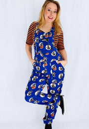 A woman modelling a pair of blue dungarees covered in a repeat print of retro spooky pin up witches, worn over a black and orange striped t shirt against a white studio background.