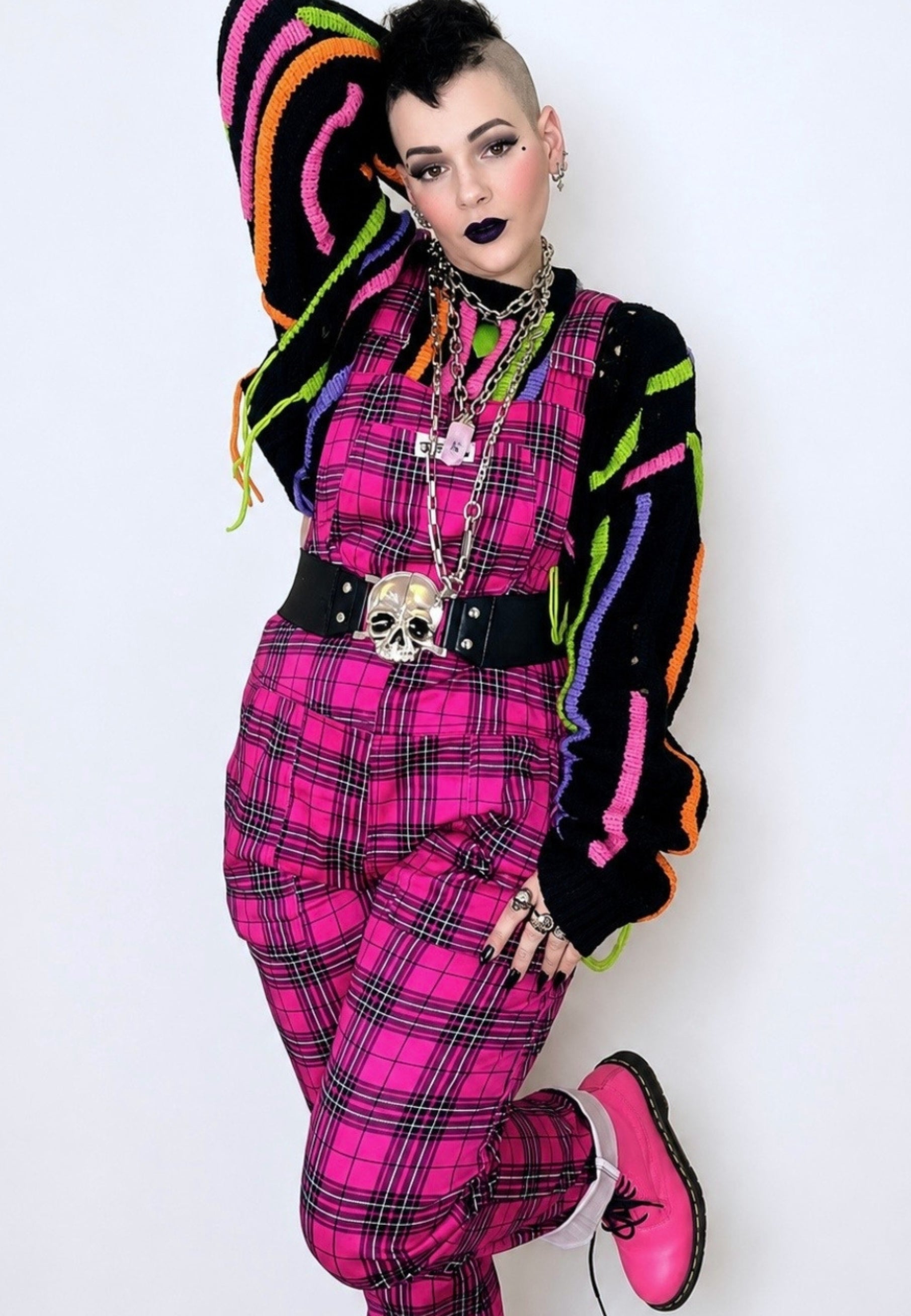 A woman wearing hot pink tartan dungarees with multiple pockets, paired with a multicolored top and matching pink boots, standing with one hand on their hip.and one hand behind their head