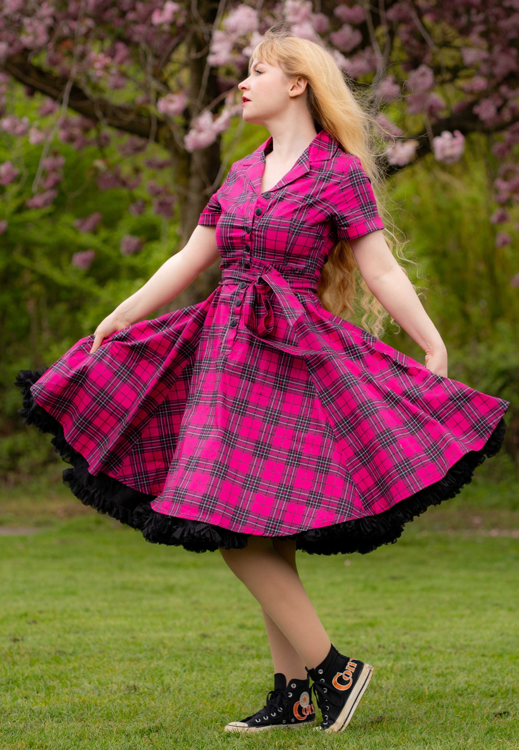 Woman in a pink tartan dress standing outdoors with cherry blossom trees in the background