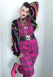 A woman wearing hot pink tartan dungarees with multiple pockets, paired with a multicolored top and matching pink boots, standing with one hand on their hip.