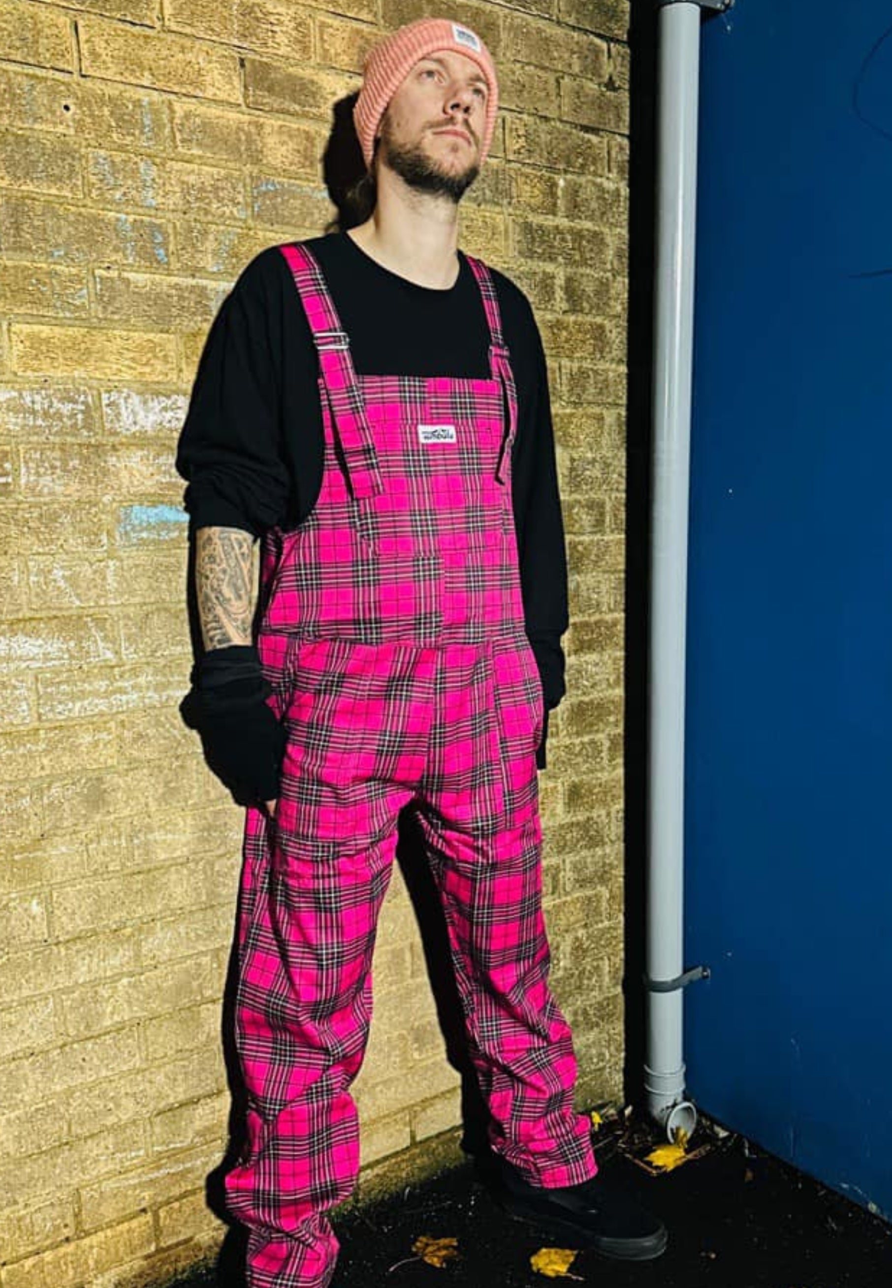 A man wearing hot pink tartan dungarees with multiple pockets, paired with a black top and  black fingerless gloves. 