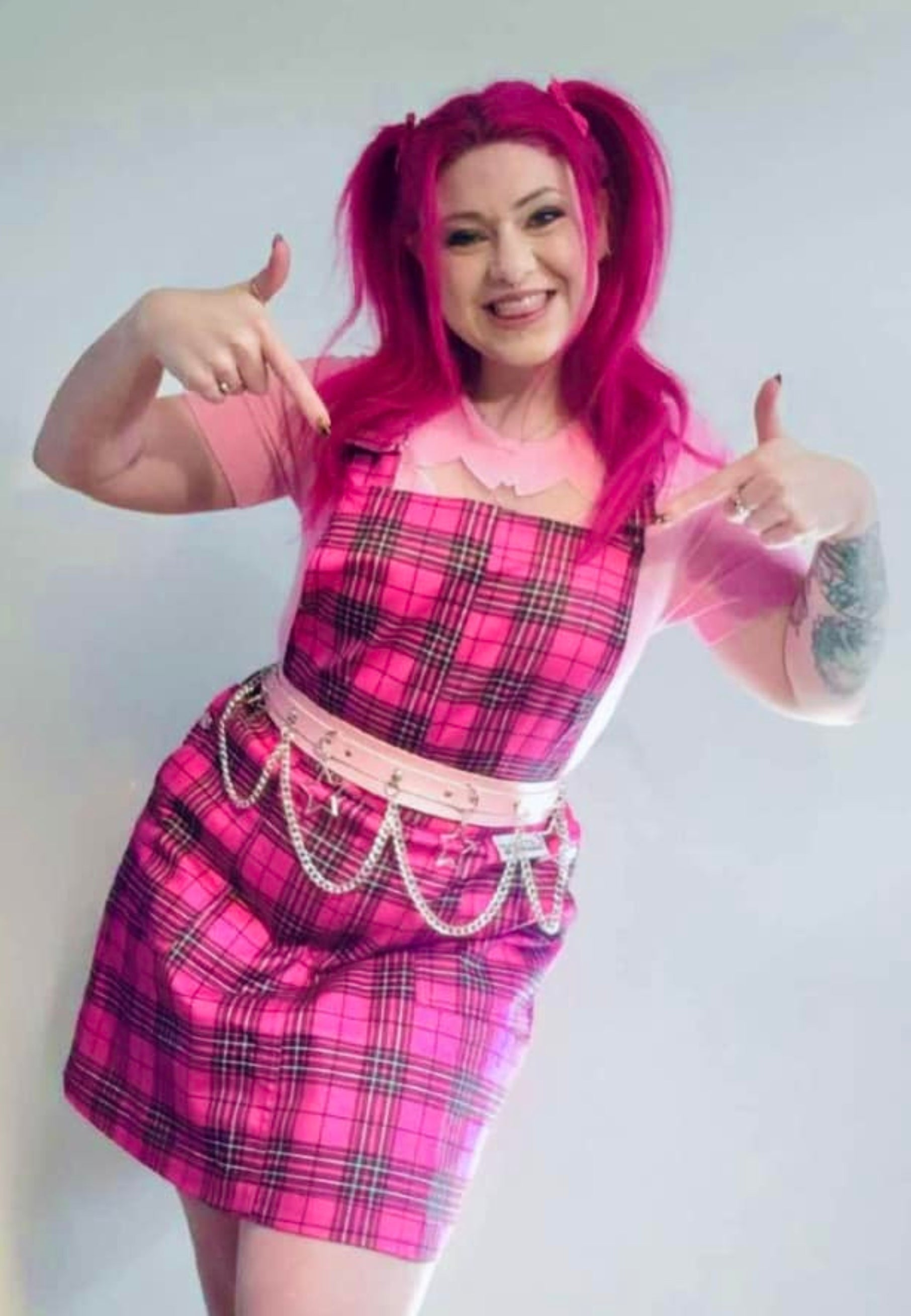 A woman with pink hair in bunches wearing a pink tartan pinafore dress with deep hip pockets and side button fastenings.