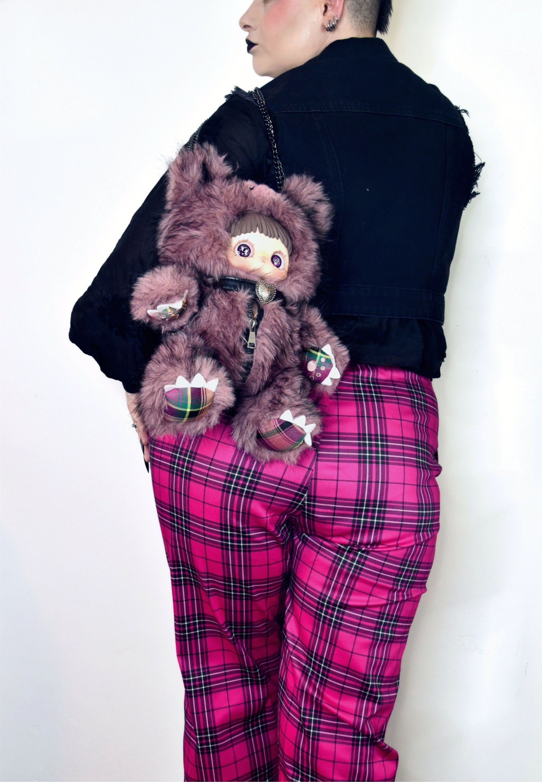 A model shown from the back wearing a black jacket and bright pink tartan trousers, carrying a brown furry doll backpack against their shoulder.