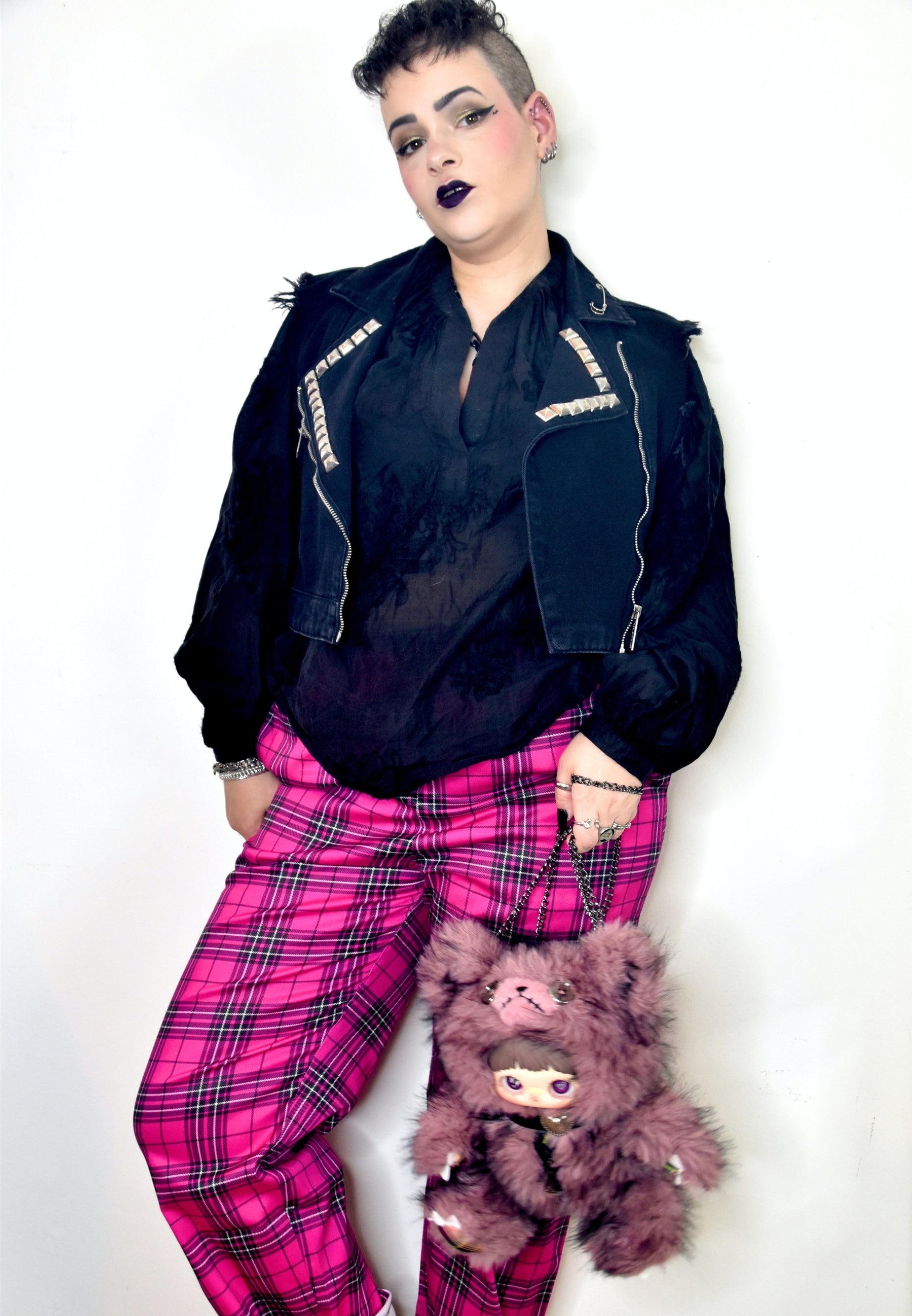 A person modeling a black denim jacket with silver studs, a sheer black top, bright pink tartan trousers, and holding a fuzzy dark pink plush bag against a plain white background.