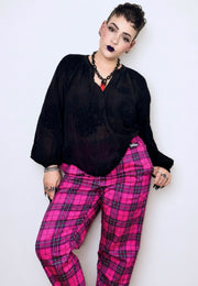 A short haired woman modeling a black sheer long-sleeve blouse and bright pink tartan trousers against a plain light background. Alternative punk goth styled 