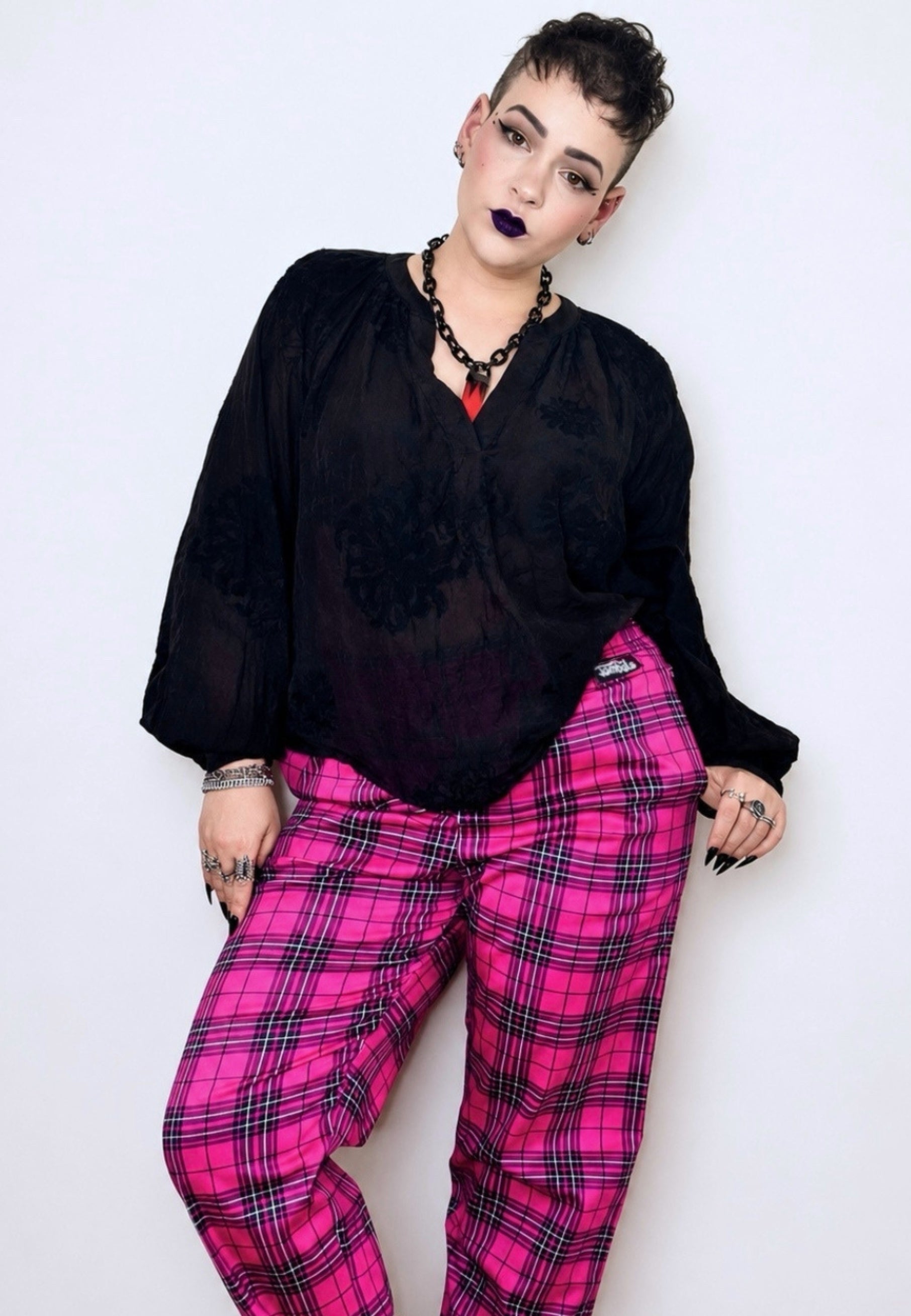 A short haired woman modeling a black sheer long-sleeve blouse and bright pink tartan trousers against a plain light background. Alternative punk goth styled 