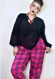 A short haired woman modeling a black sheer long-sleeve blouse and bright pink tartan trousers against a plain light background. Alternative punk goth styled 