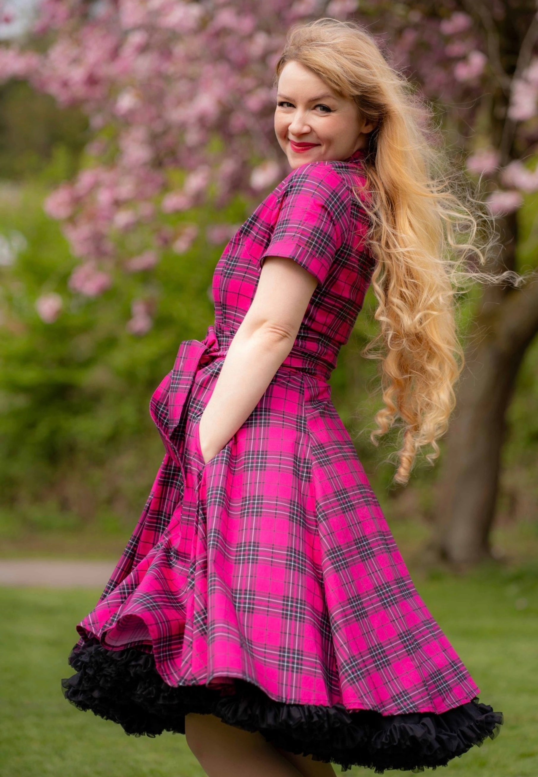 Woman in a pink tartan dress with black ruffles standing in a garden with cherry blossoms.