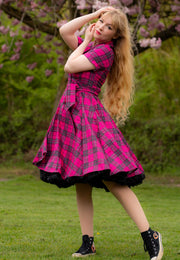 Woman in a pink tartan dress standing outdoors with greenery and cherry blossom in the background