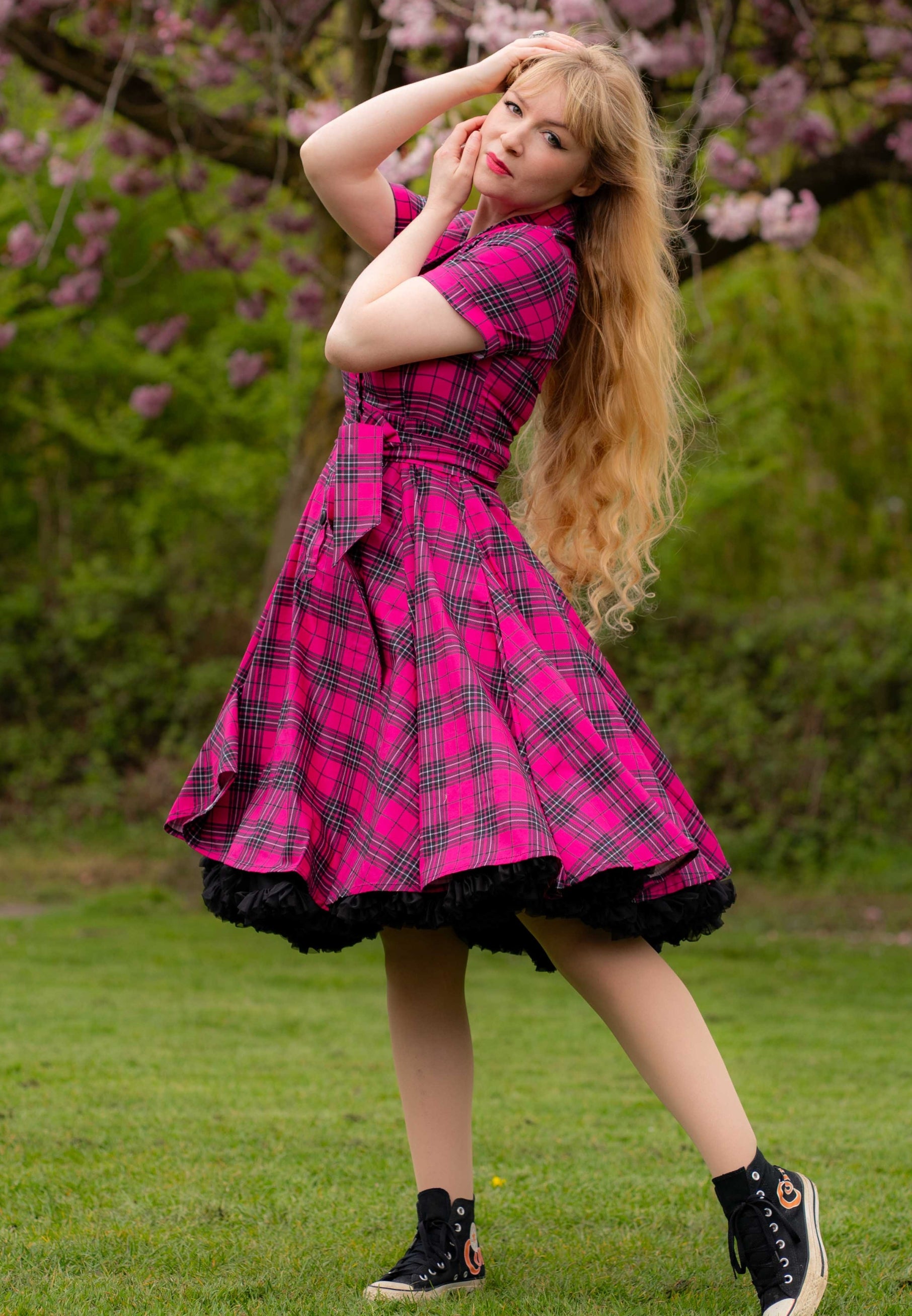 Woman in a pink tartan dress standing outdoors with greenery and cherry blossom in the background