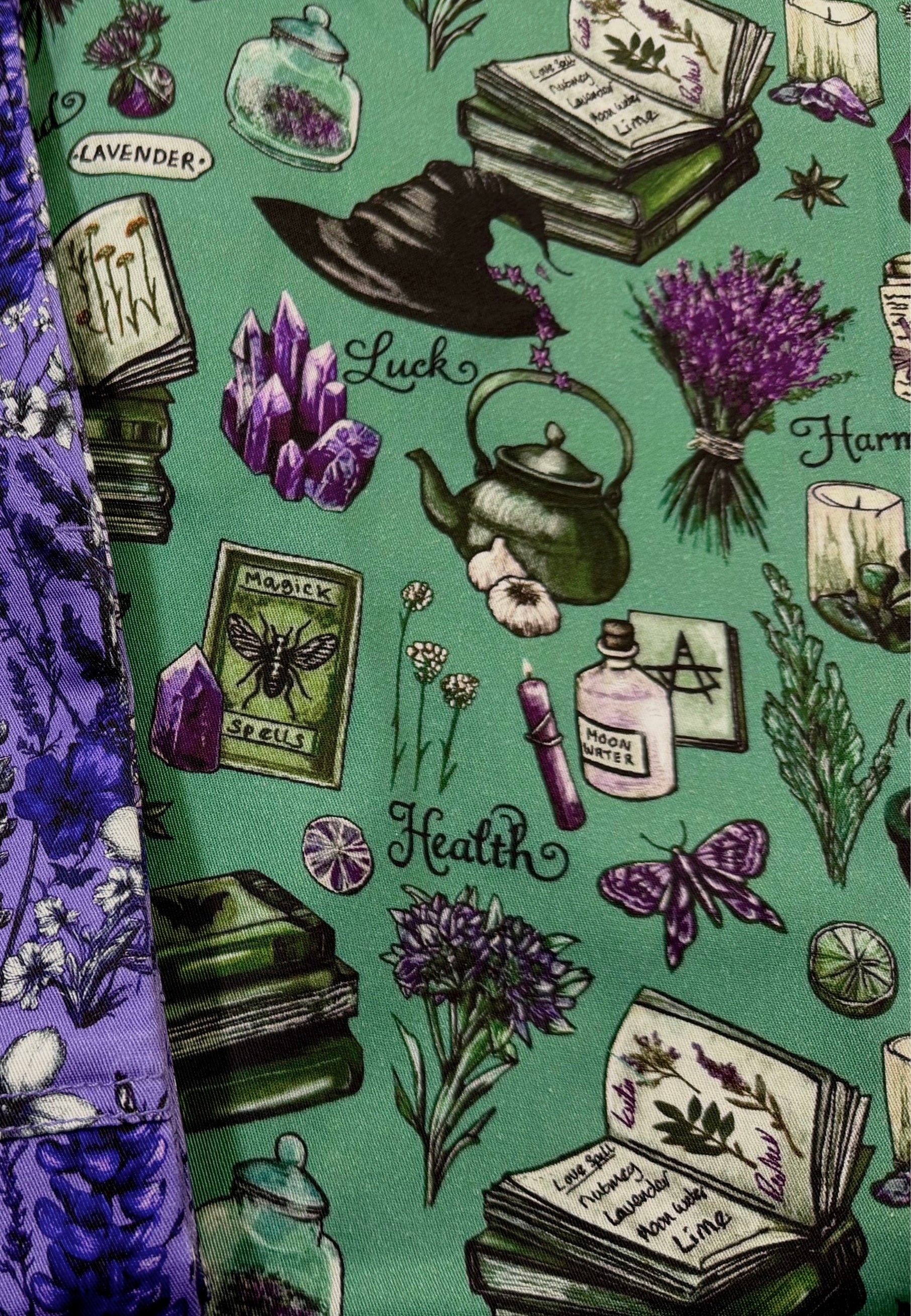 A close up of a fabric pront featuring a witch themed design on a sage green background, with illustrations of books, a cauldron, crystals, lavender, a witch hat, a potion bottle, and the words 'Luck' and 'Health'. 