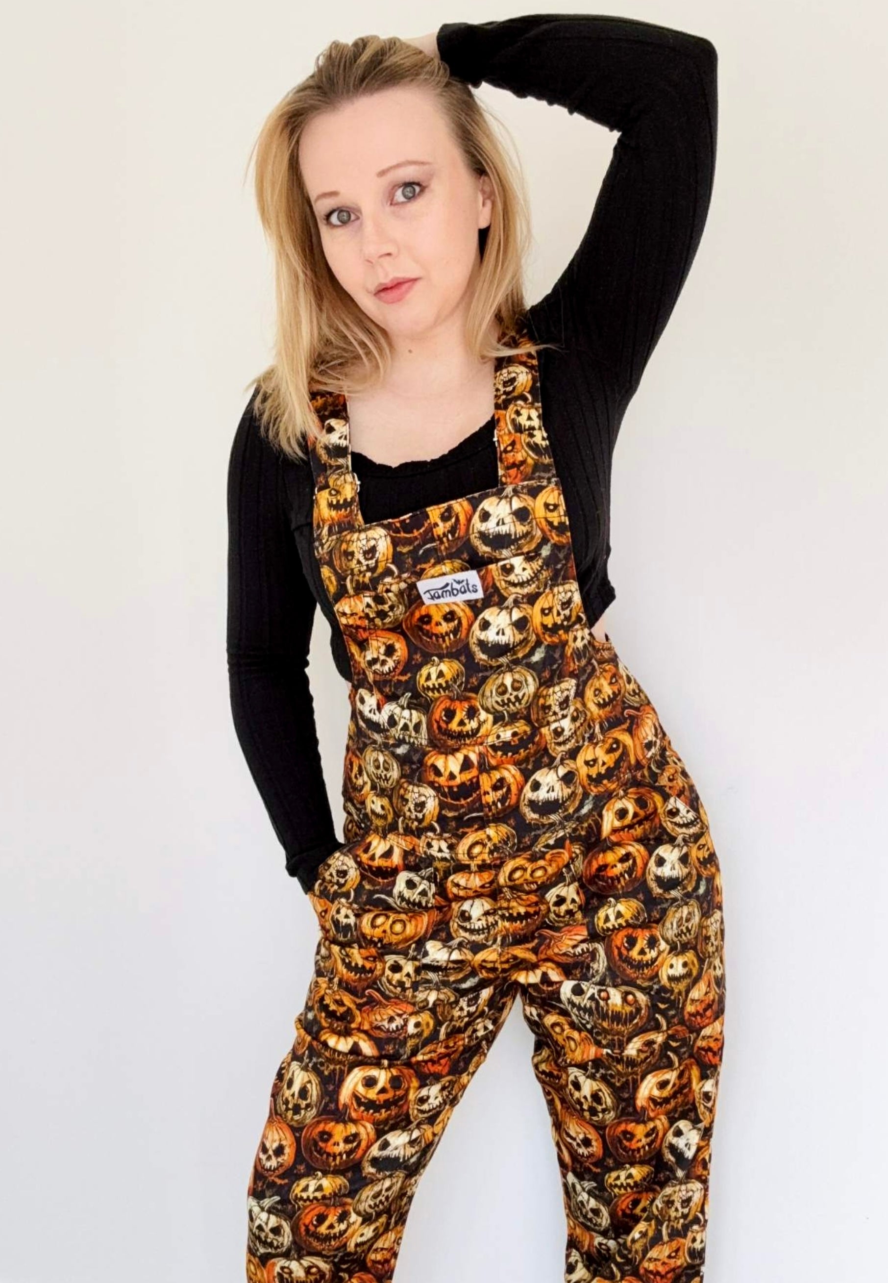 Model wearing Halloween themed dungarees overalls with illustrated creepy pumpkins print. Model is also wearing long sleeved black top. Studio photo on a white background