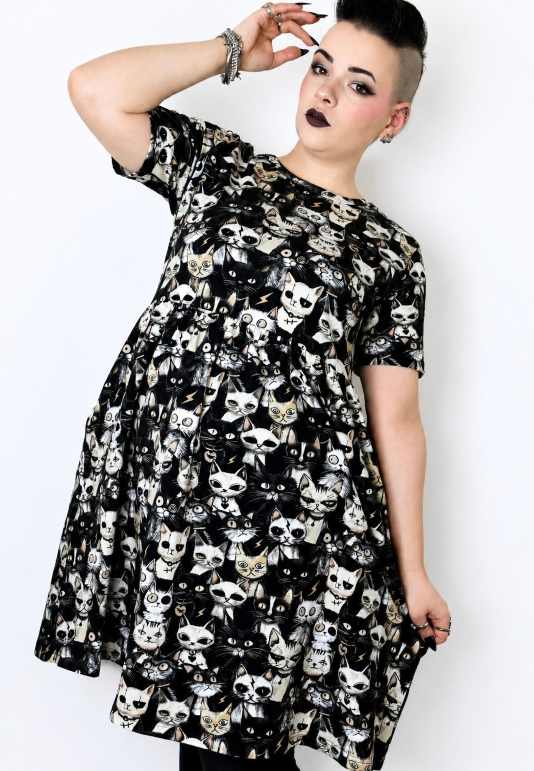 A woman modelling a black smock dress covered in a repeating white and beige punky alley cat print against a plain light background.