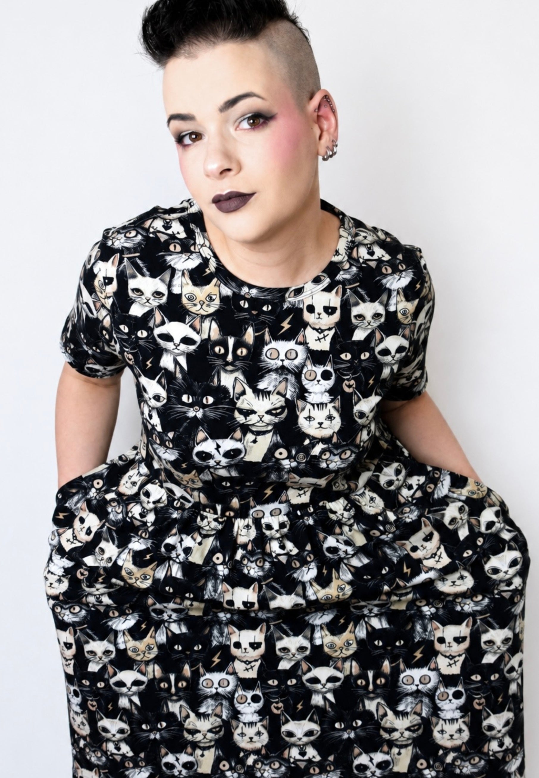 A person with a short dark mohawk hairstyle wearing a black smock dress covered in a white and grey punky cat pattern, posed against a plain background. model is holding hands in pockets to show off the pockets. 