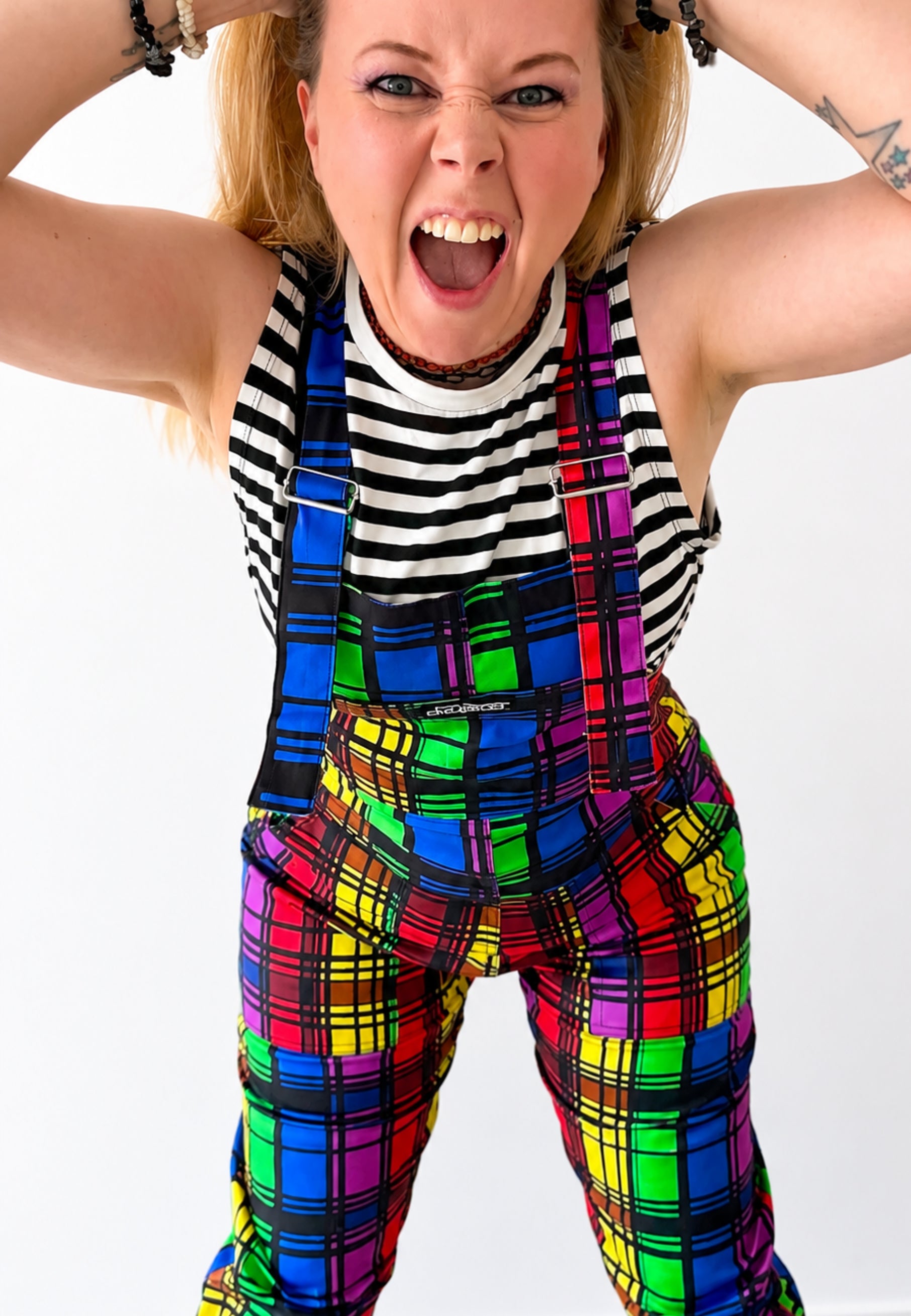 Woman wearing a bright coloured rainbow tartan dungarees with a black and white striped top and big boots. Woman is standing against a white background and she has her hands on her head shouting 