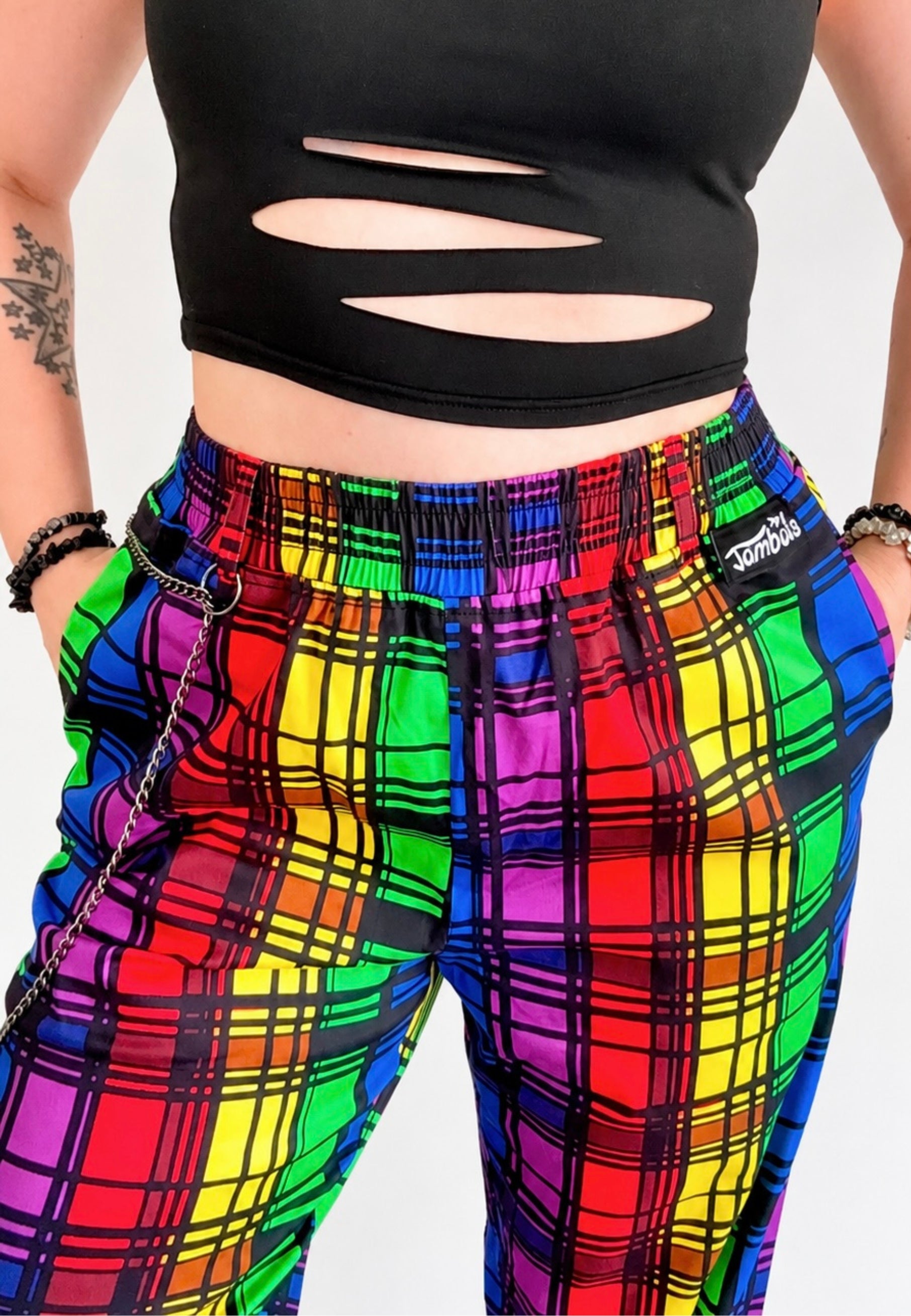 Person wearing a black ripped crop top and vibrant rainbow tartan trousers with a punky chain on belt looks. Photo is on a plain background