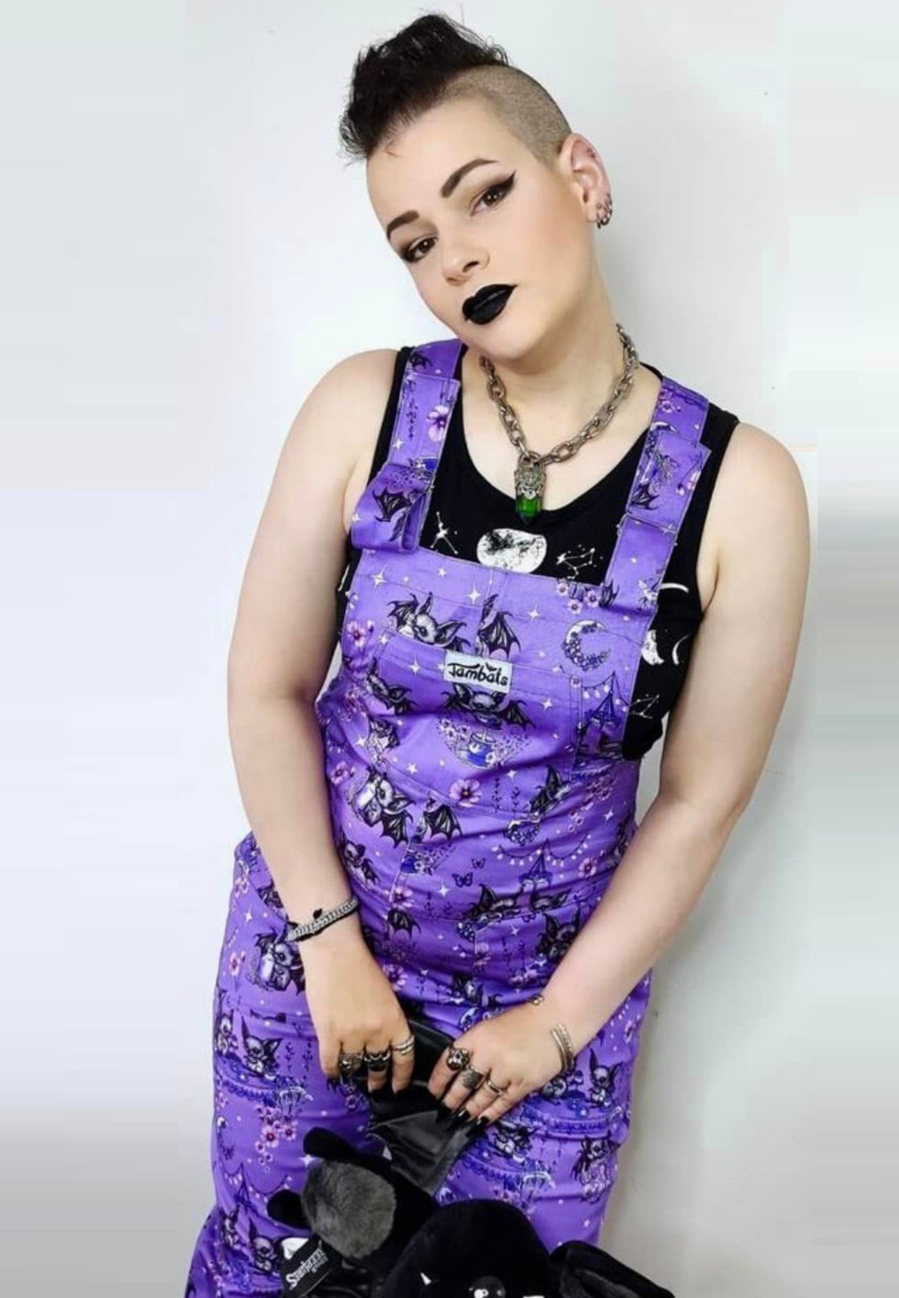 A woman wearing a deep lavender/soft purple dungarees with a cute gothic bats tea party print, paired with a black top. Cute alternative style. 