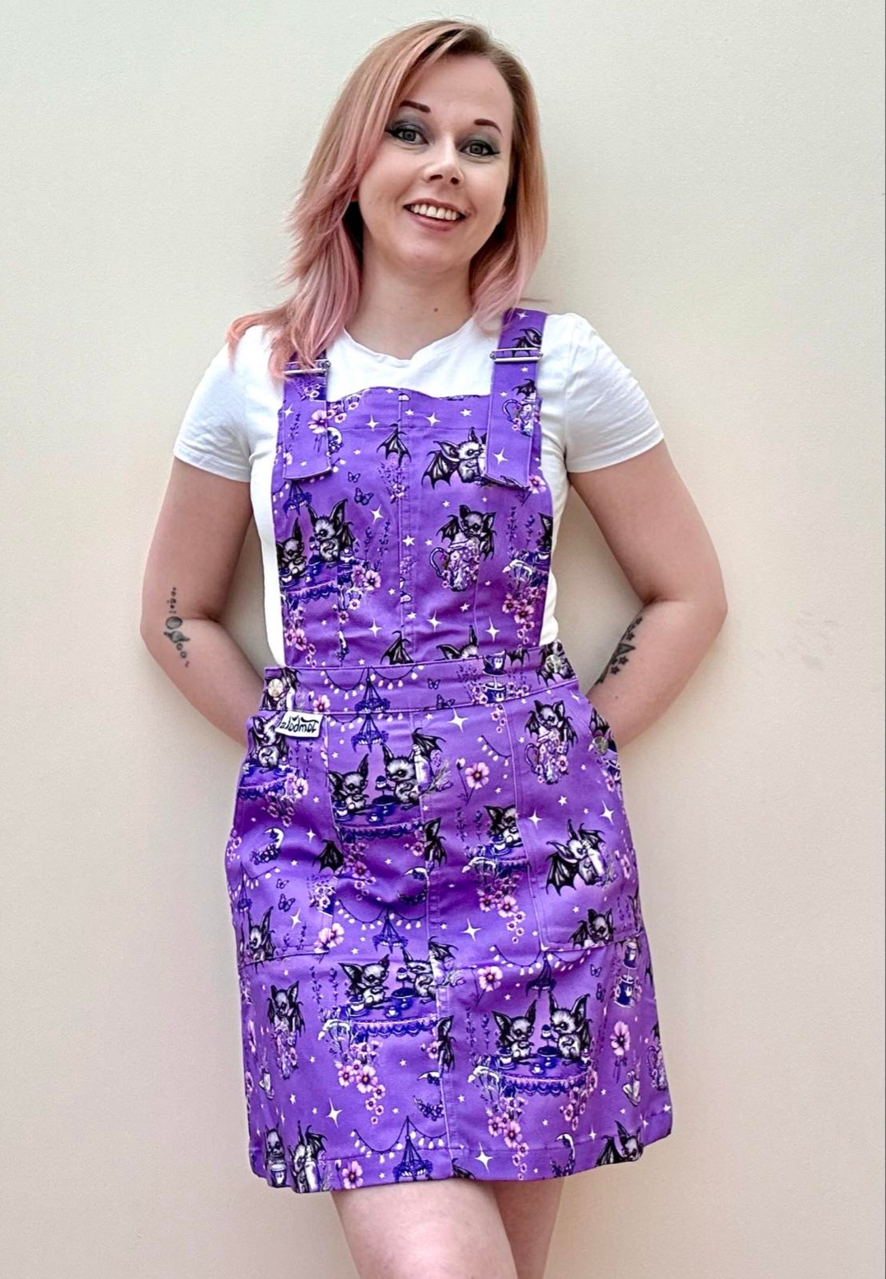 A woman wearing a deep lavender purple pinafore dress with a cute and whimsical bats tea party print, over a white t-shirt, standing against a neutral background. Cute goth print pinafore dress. 