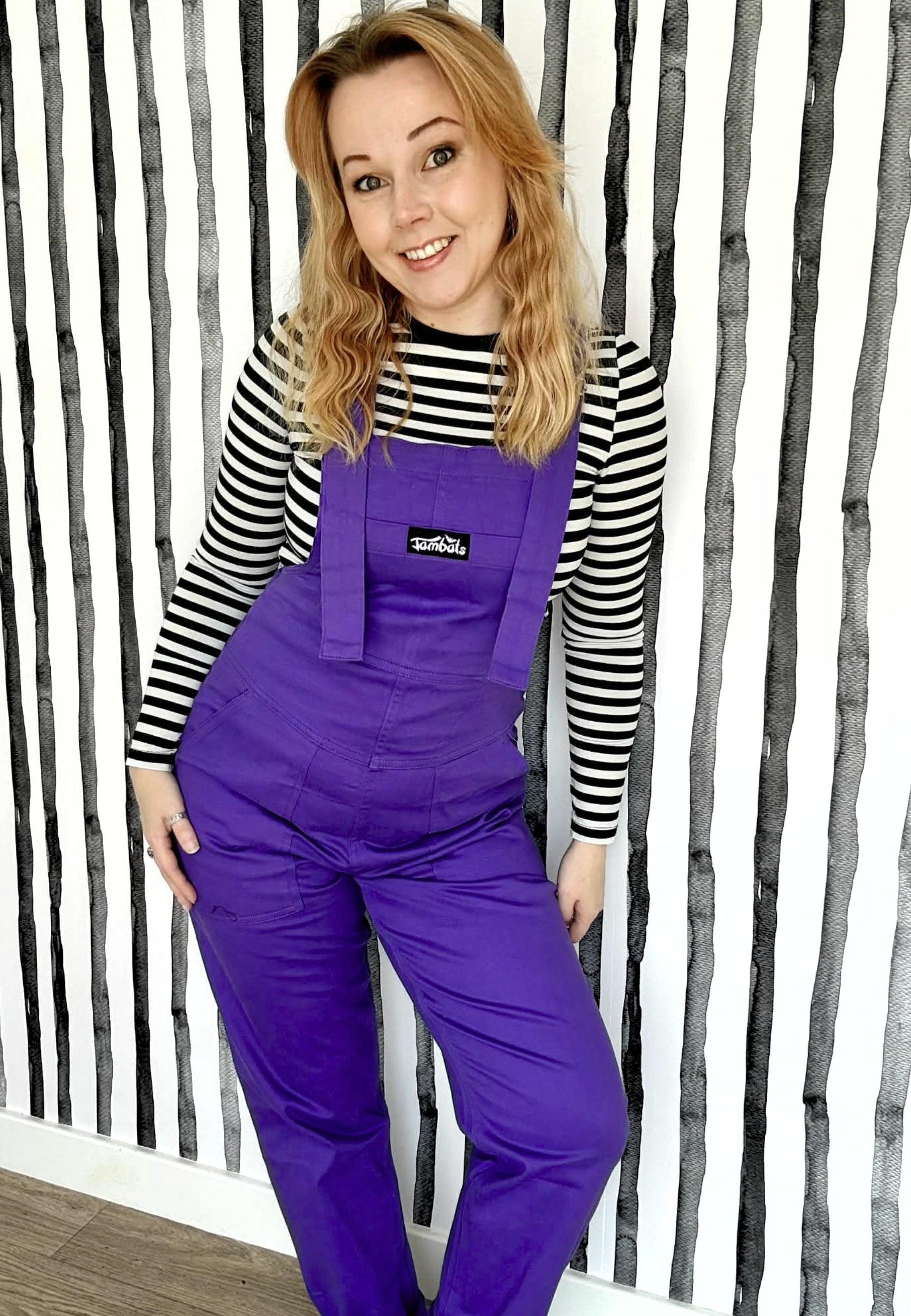Woman wearing purple dungarees with a striped shirt against a striped wall background