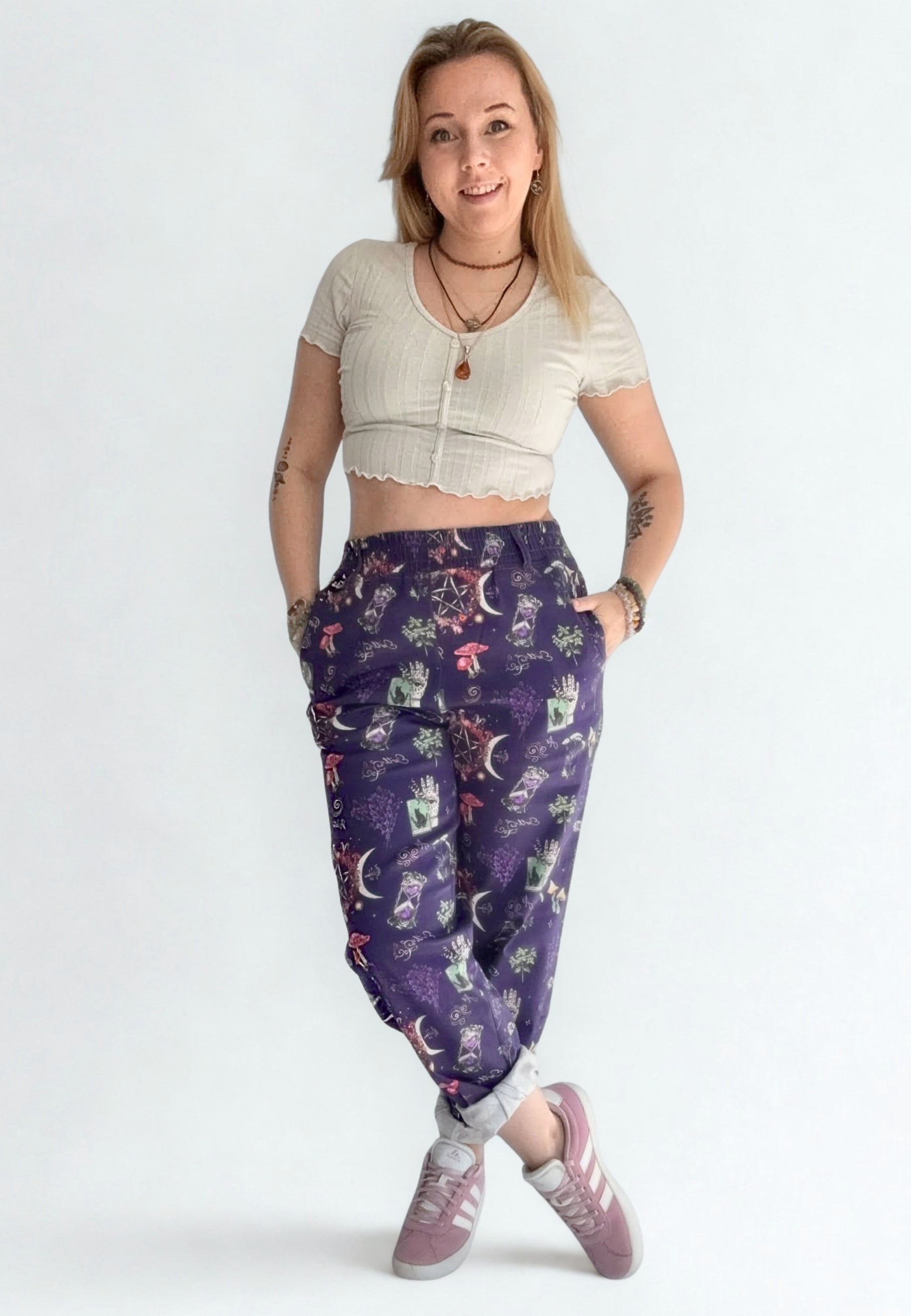 Woman wearing a beige crop top and purple mystical patterned trousers and Vans pumps on a white background