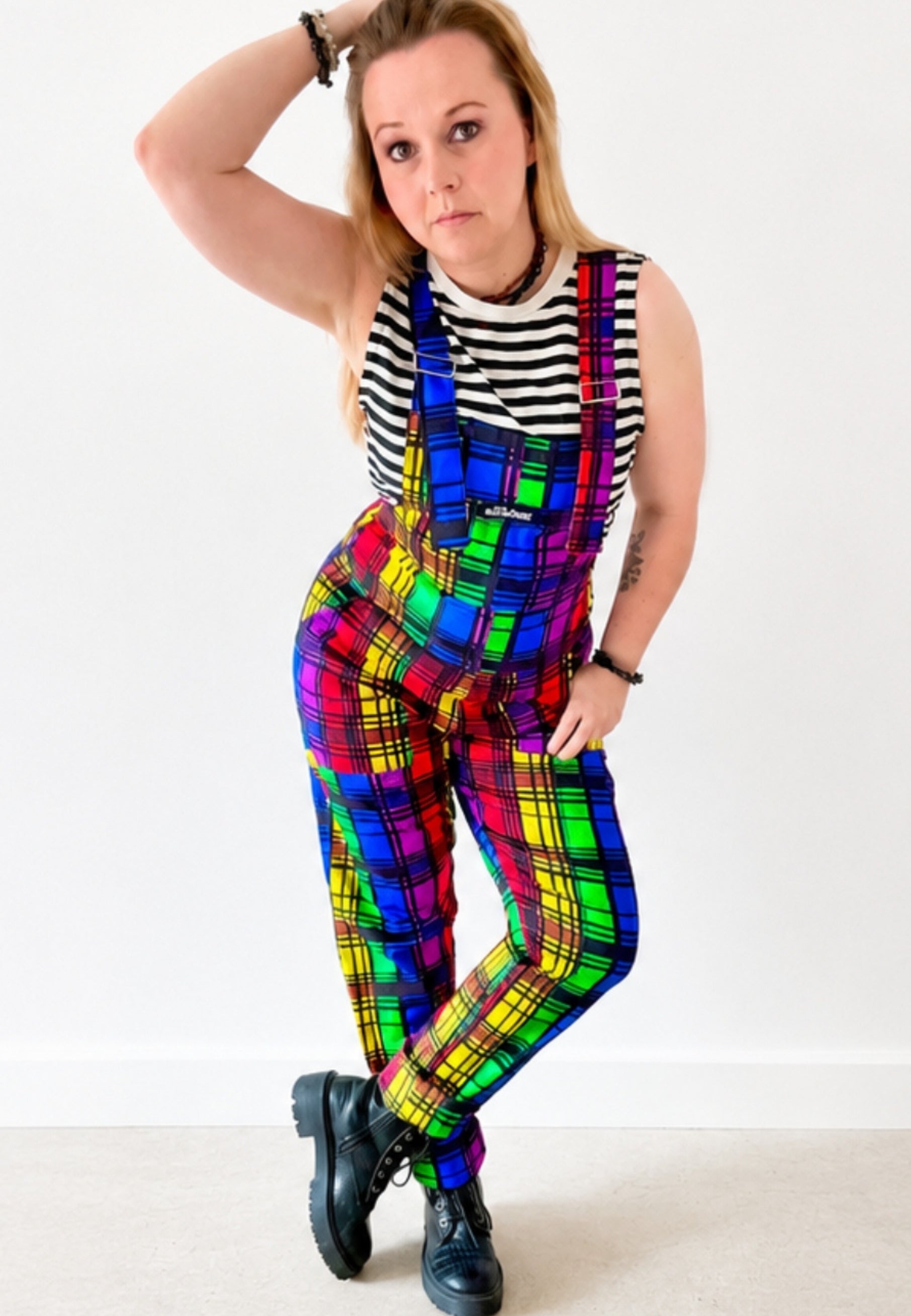 Woman wearing a bright coloured rainbow tartan dungarees with a black and white striped top and big boots. Woman is standing against a white background