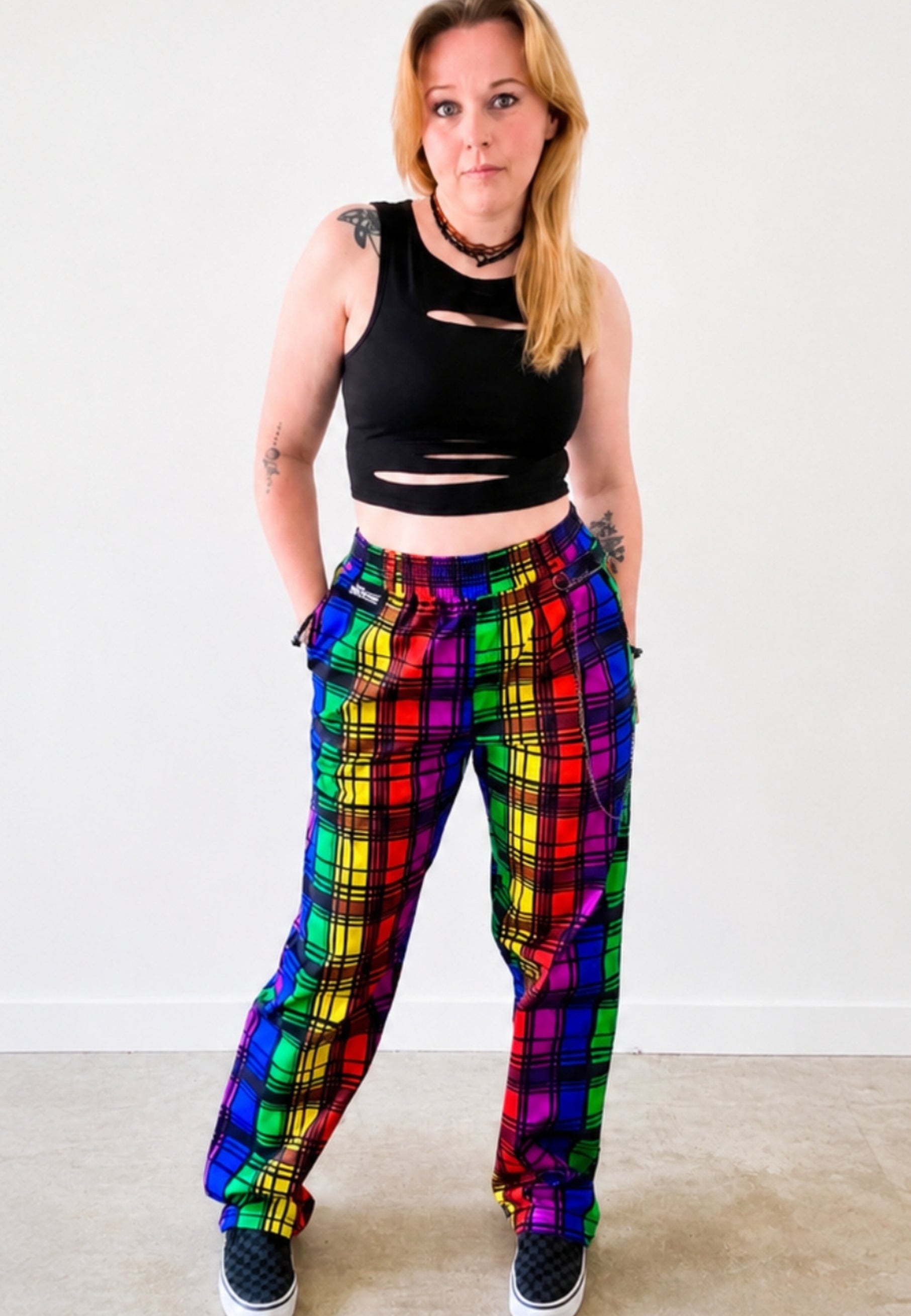 Person wearing a black ripped crop top and vibrant rainbow tartan trousers with a punky chain on belt looks. Photo is on a plain background