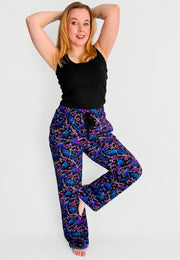 A woman modelling a pair of wide leg lounge pants with a vibrant purple and blue soft gothic raven and crow print , paired with a black tank top, against a plain light gray background