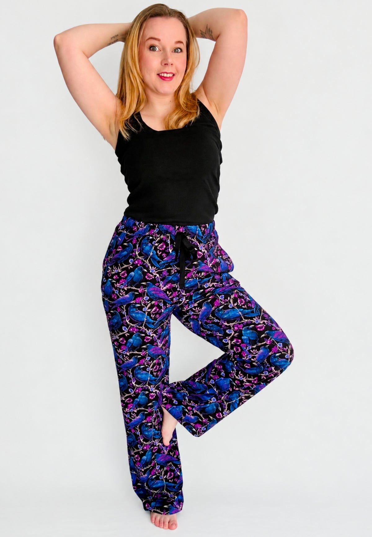 A woman modelling a pair of wide leg lounge pants with a vibrant purple and blue soft gothic raven and crow print , paired with a black tank top, against a plain light gray background