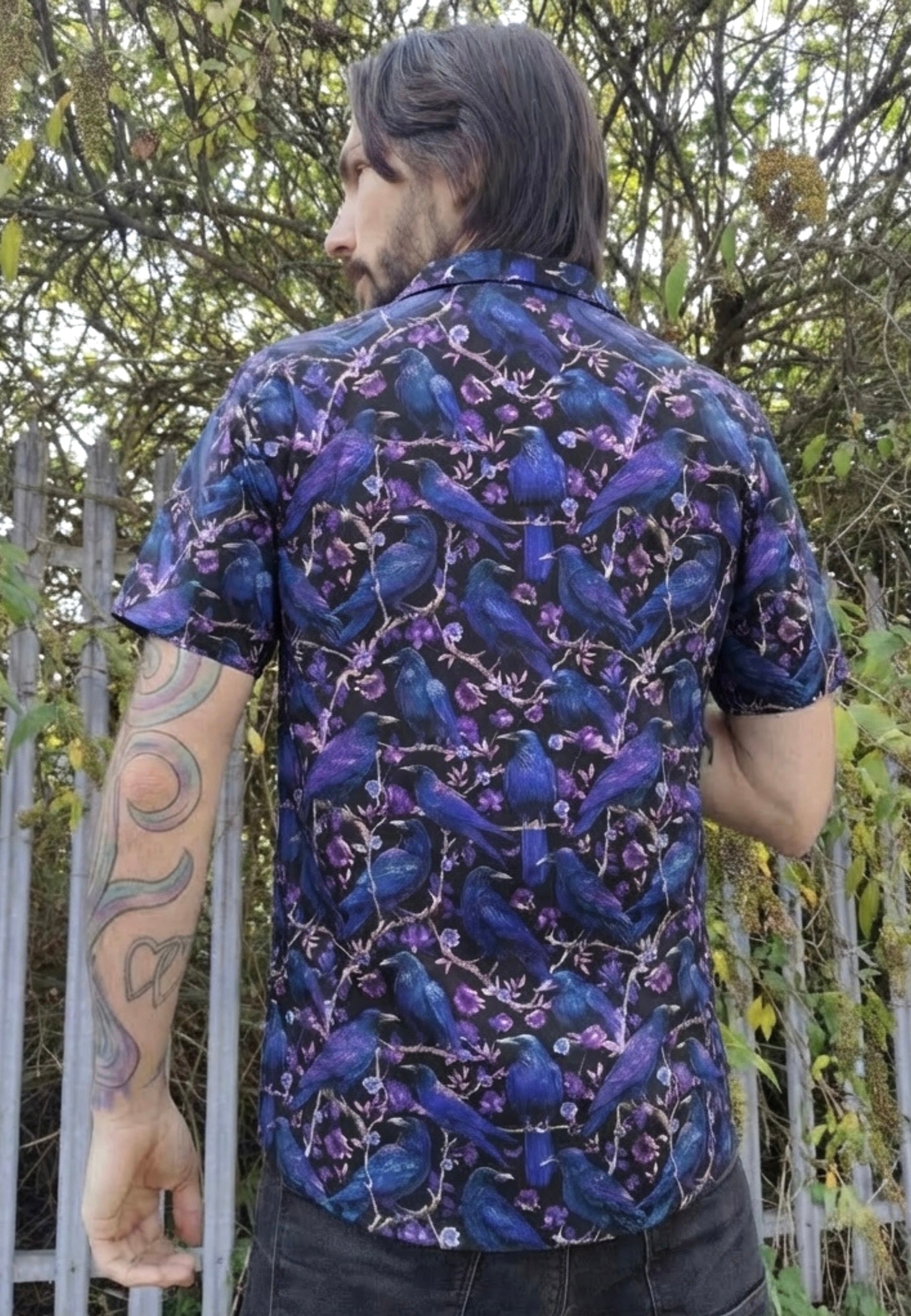 Man wearing a shirt with a print of Corvids (crows and ravens). Trees and railings in the background