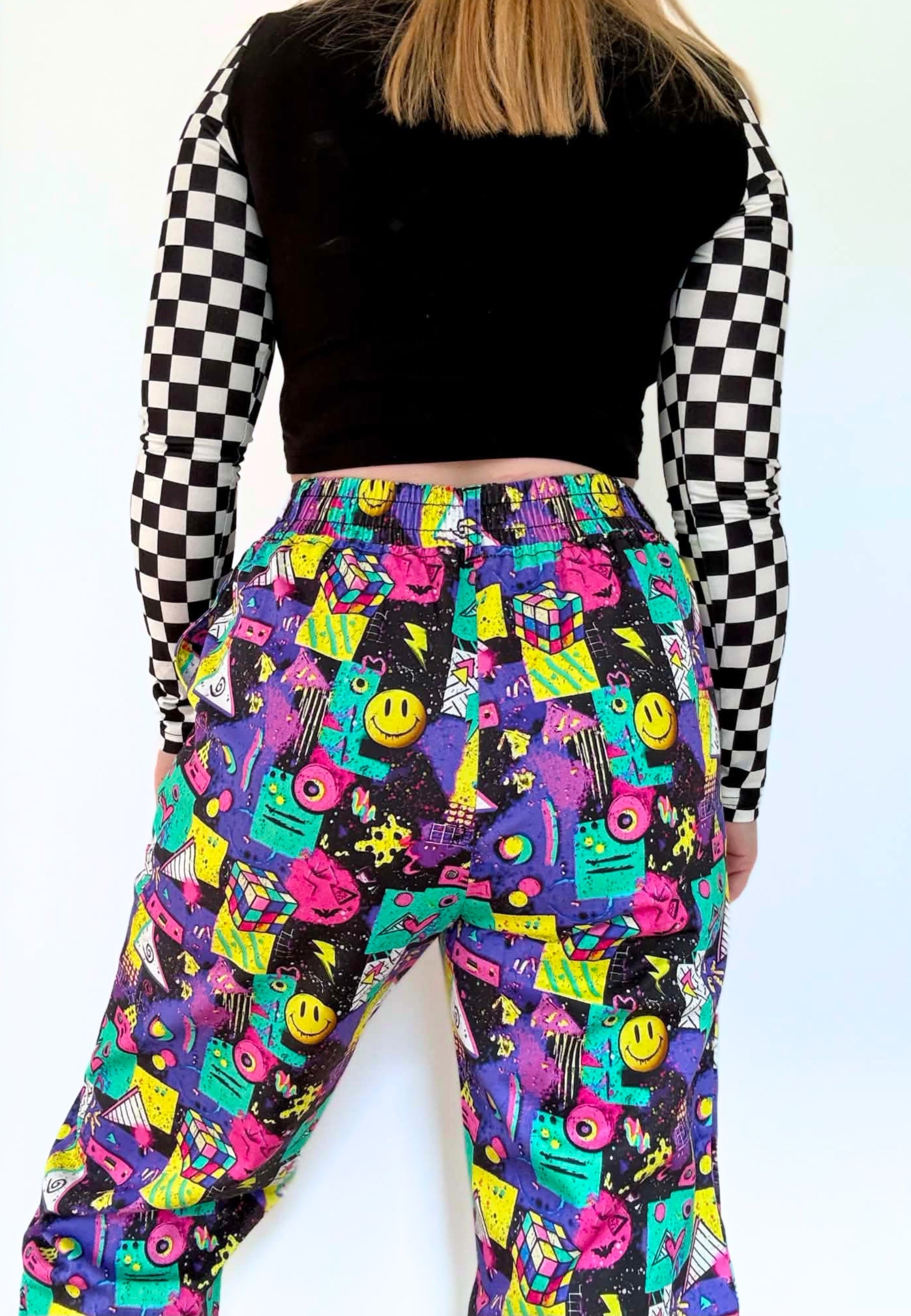 A person shown from the back wearing colourful, 80s 90s patterned wide leg pants with a black and white checkered long-sleeve top. The pants feature a vibrant, multicolored retro graphic print with smiley faces, stars, and geometric shapes on a black background.