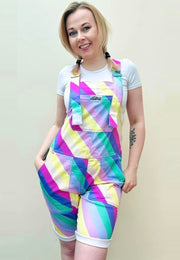 A person wearing bright multicolored striped shorts dungarees with a chest pocket, standing against a plain background. Bright pastel striped dungaree shorts. Retro style. 