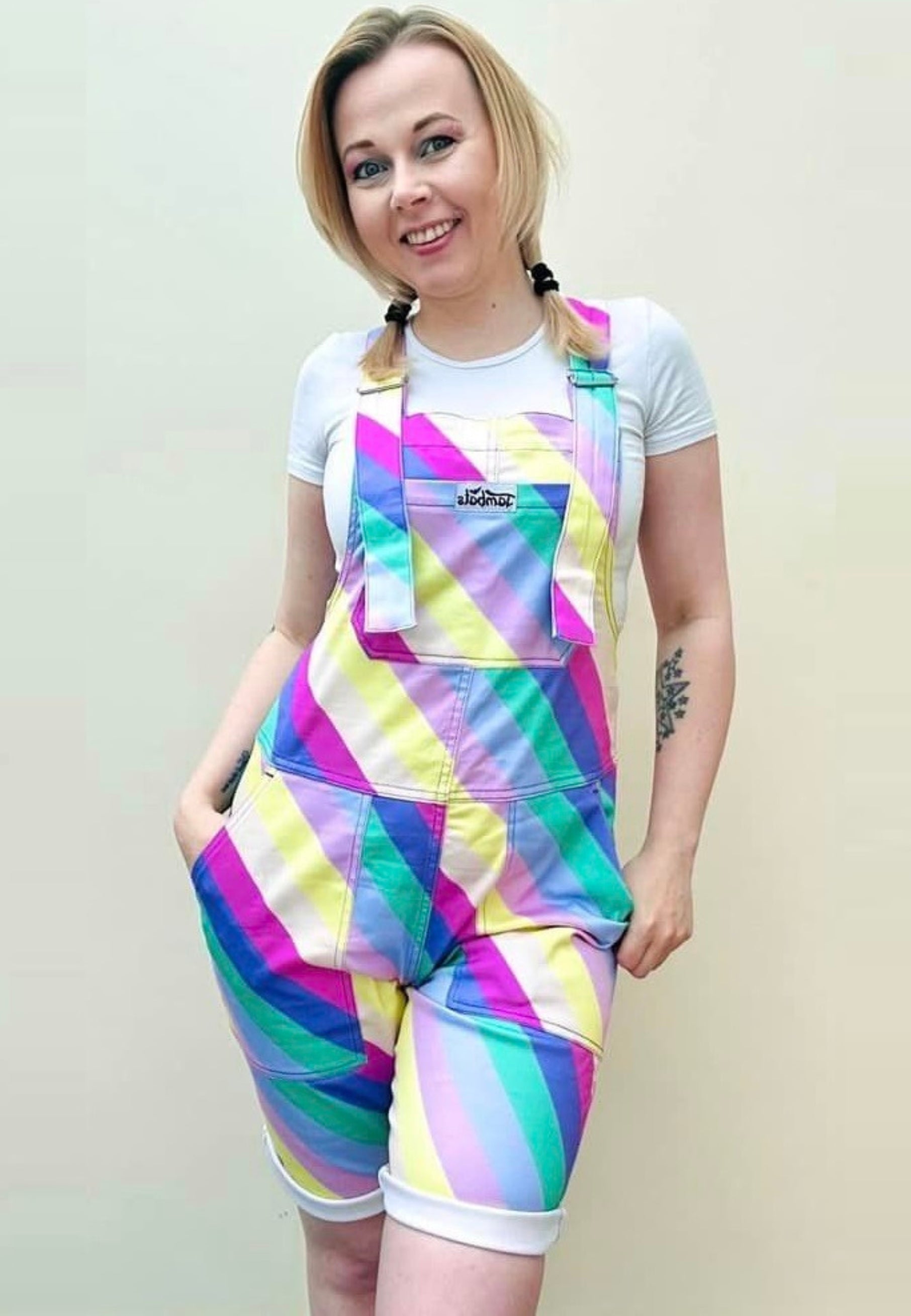 A person wearing bright multicolored striped shorts dungarees with a chest pocket, standing against a plain background. Bright pastel striped dungaree shorts. Retro style. 