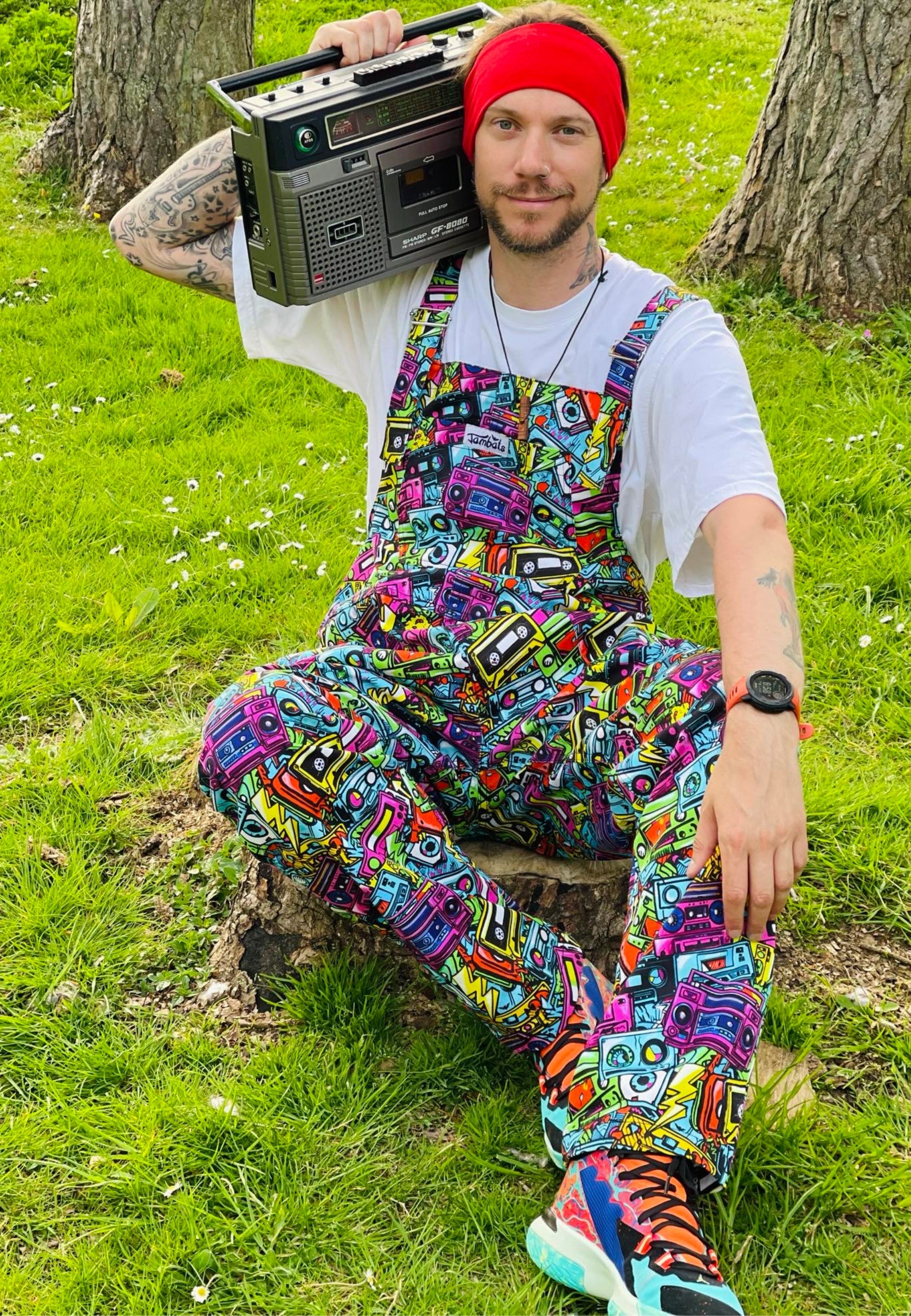 A man wearing a colourful, retro styled  outfit with a print on the dungarees of retro cassette tapes in a street art style. Man is holding a big retro ghetto blaster 