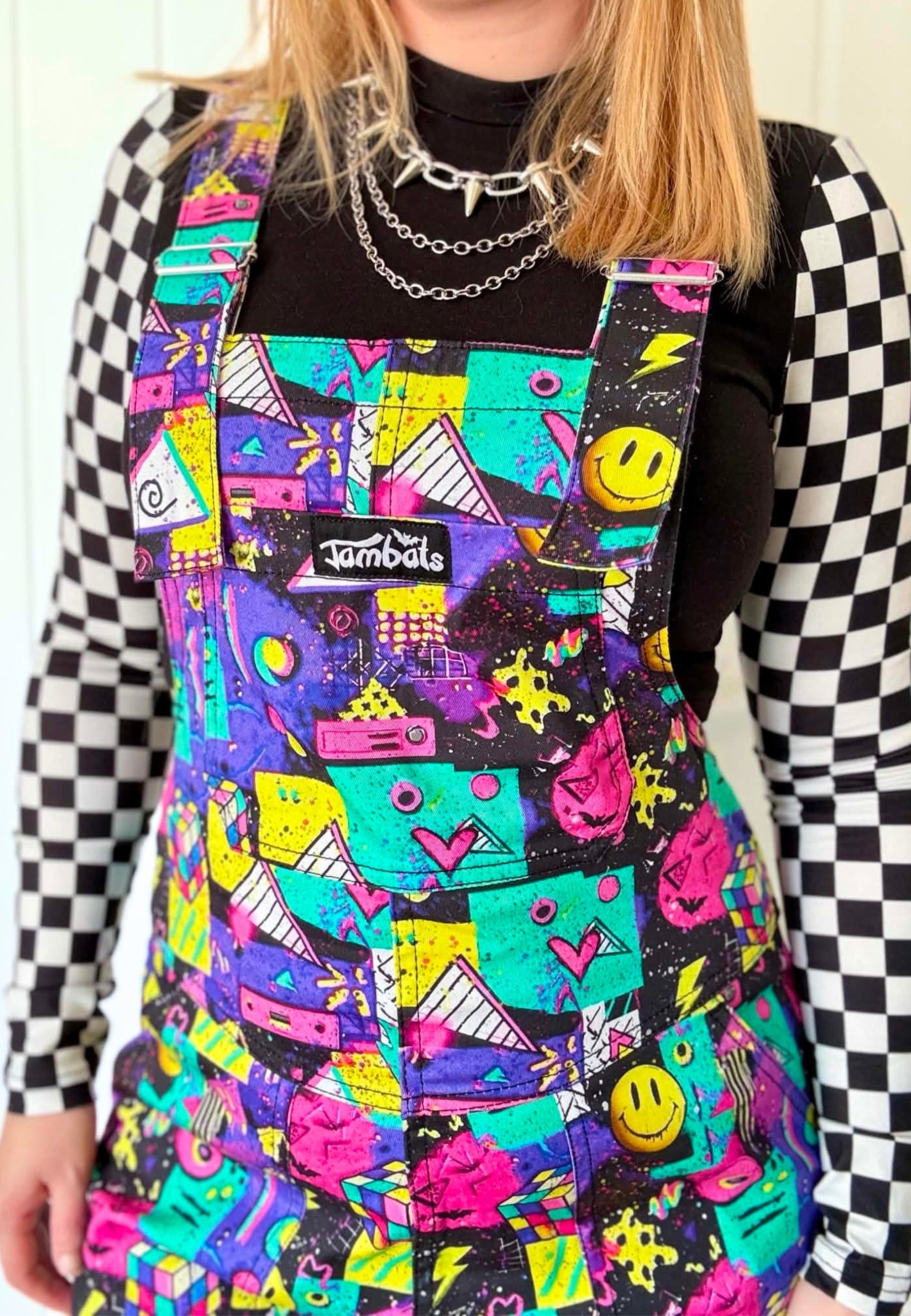 A person wearing a colourful 80s 90s print dungarees with a black and white checkered lon sleeve shirt underneath. The apron features a vibrant multicolour pattern of pink, yellow, green, and purple graphics including smiley faces, guitars, and hearts, with a visible Jambats brand label on the chest pocket.