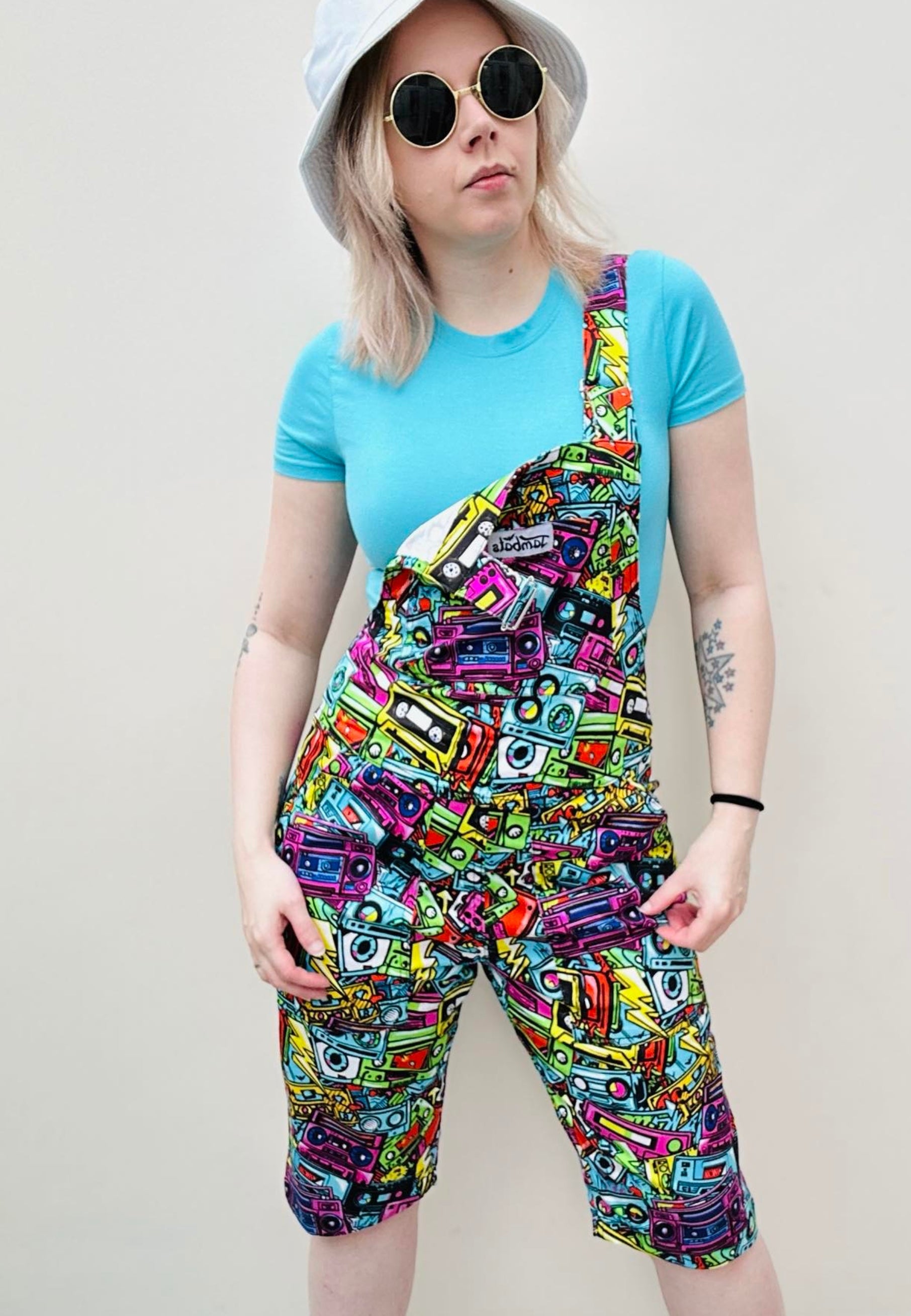 A woman wearing a pair of colourful, patterned shorts dungarees with multiple pockets and a blue t-shirt, accessorised with a white hat and sunglasses. Dungaree Shorts print is of retro graffiti style music cassettes 