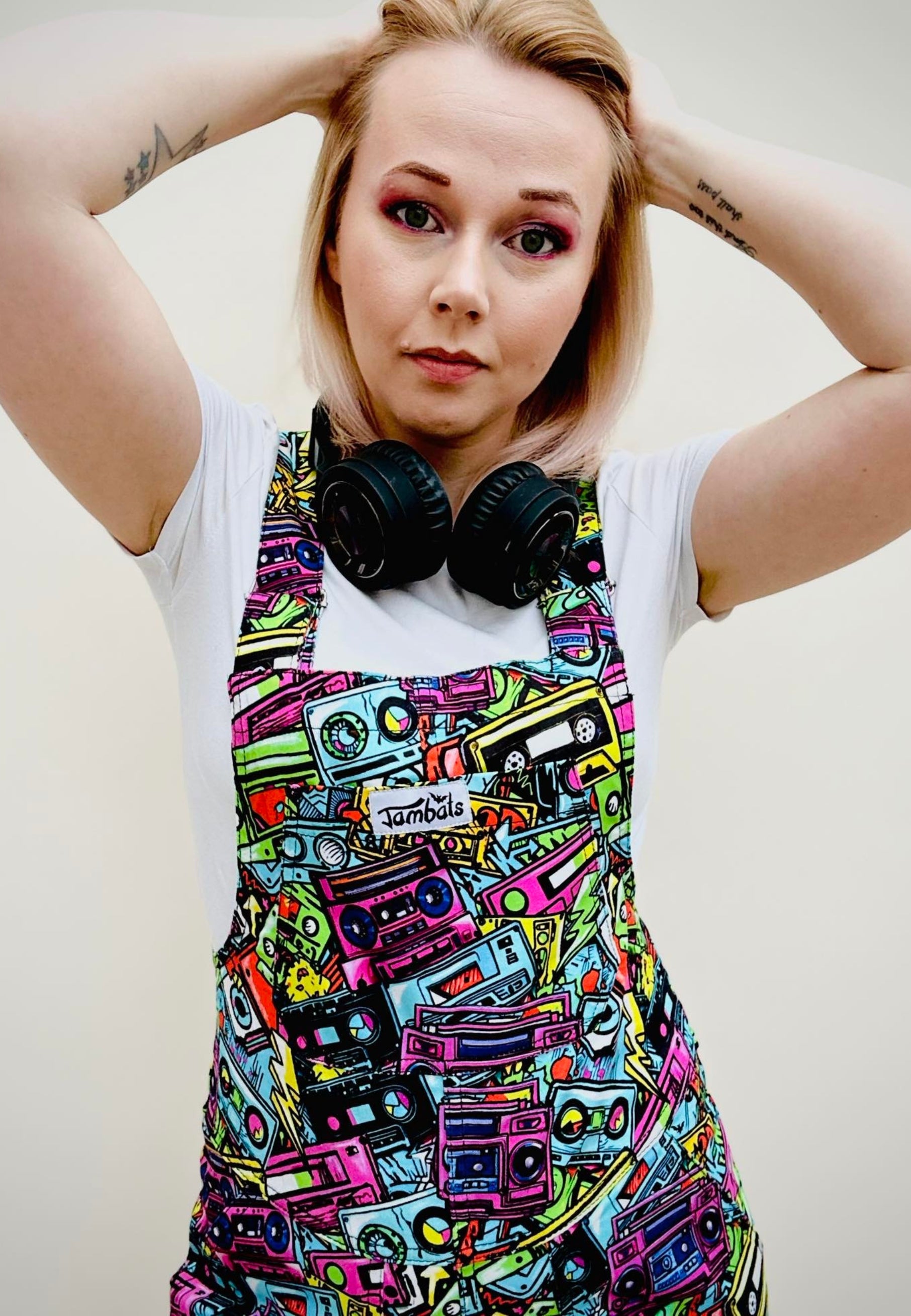 A woman wearing a pair of colourful, patterned shorts dungarees with multiple pockets and a blue t-shirt, accessorised with a big earphones. Dungaree Shorts print is of retro graffiti style music cassettes 