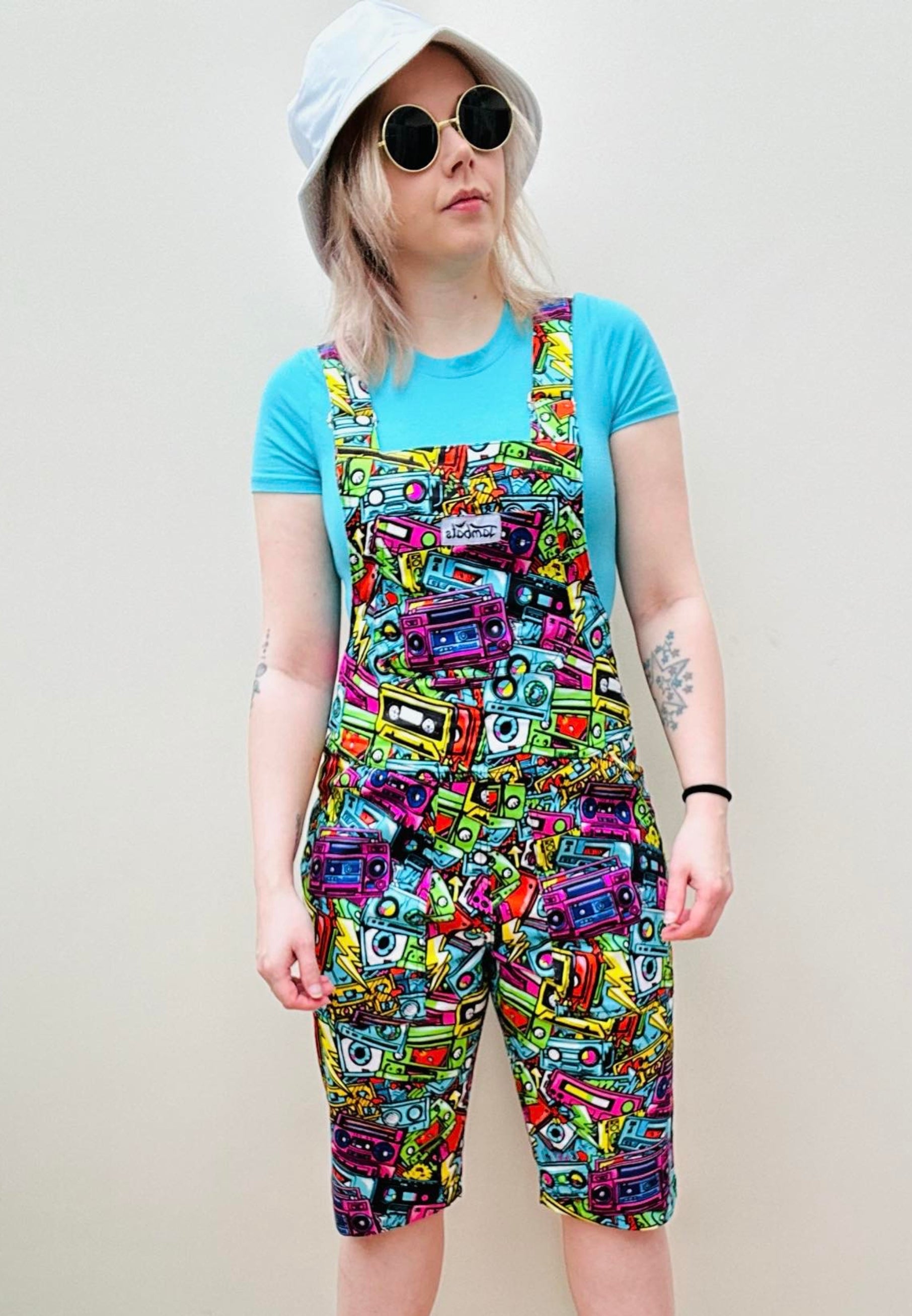 A woman wearing a pair of colourful, patterned shorts dungarees with multiple pockets and a blue t-shirt, accessorised with a white hat and sunglasses. Dungaree Shorts print is of retro graffiti style music cassettes 