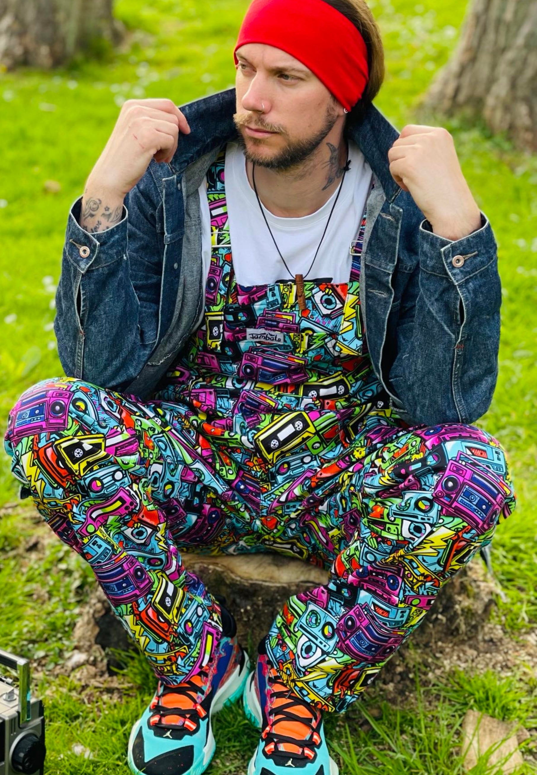 A man wearing a colourful, retro styled  outfit with a print on the dungarees of retro cassette tapes in a street art style.
